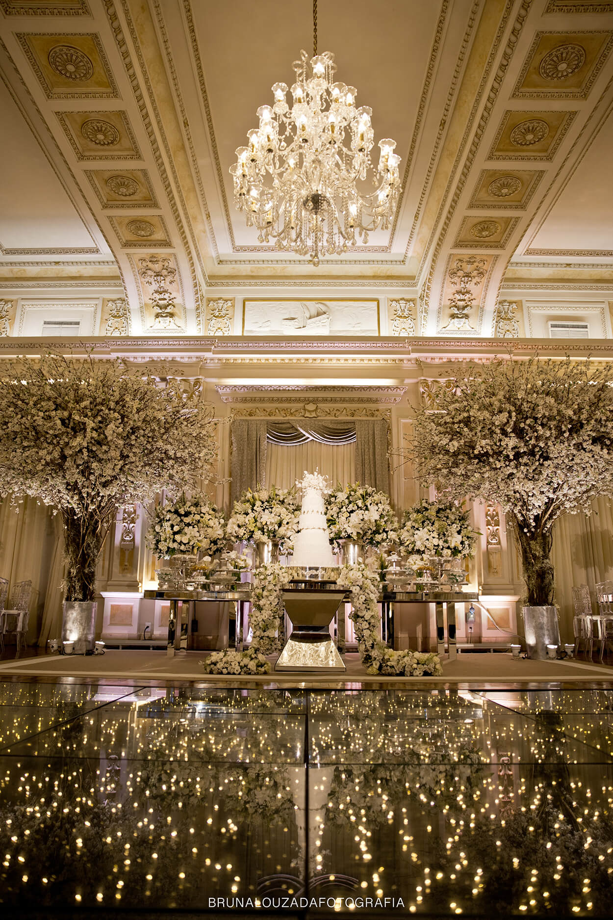 Mesa com bolo de casamento com arranjos brancos em salão grandioso e antigo com lustre