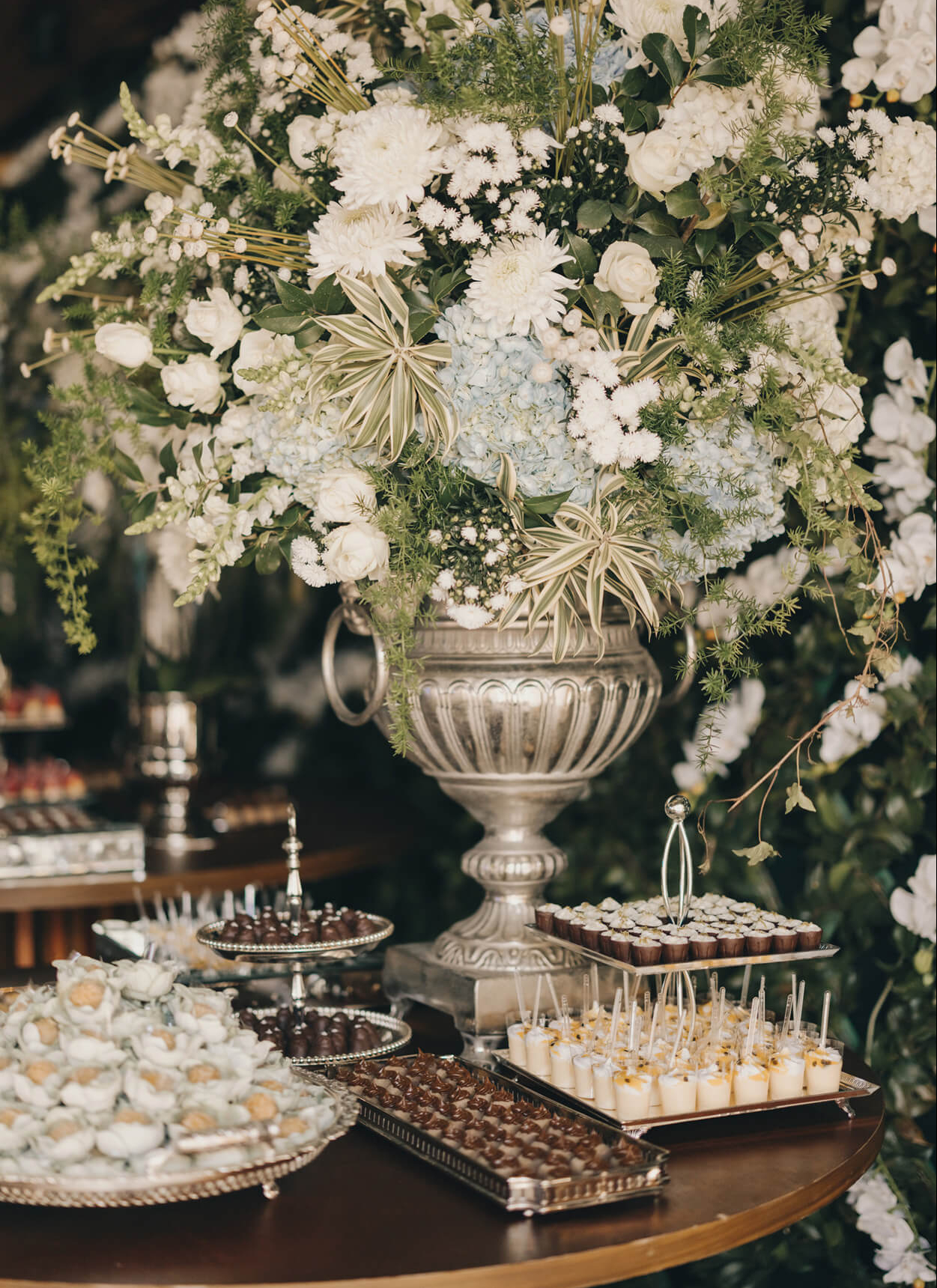 Vaso prateado com flores brancas e pratos com doces de casamento na mesa