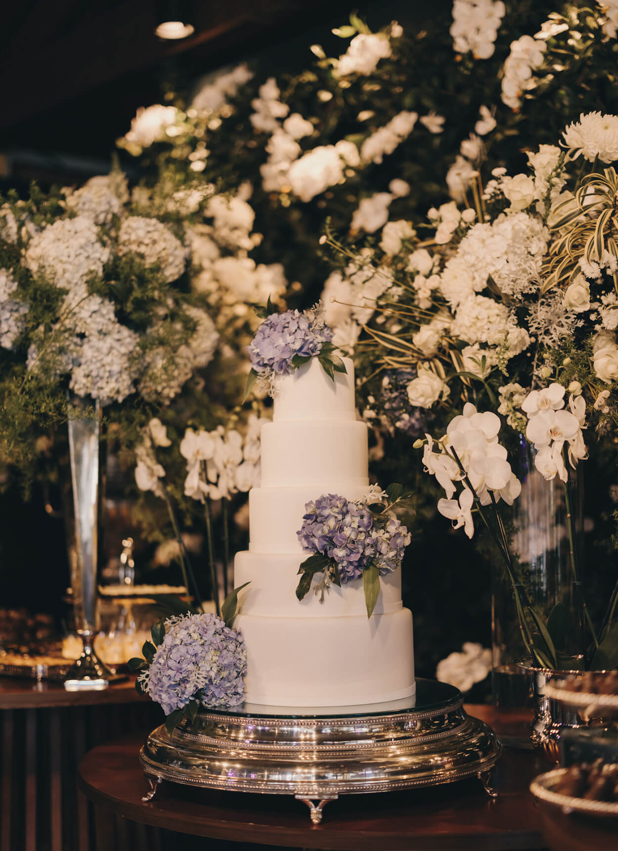 Mesa com bolo de casamento branco com quatro andares e flores lilás