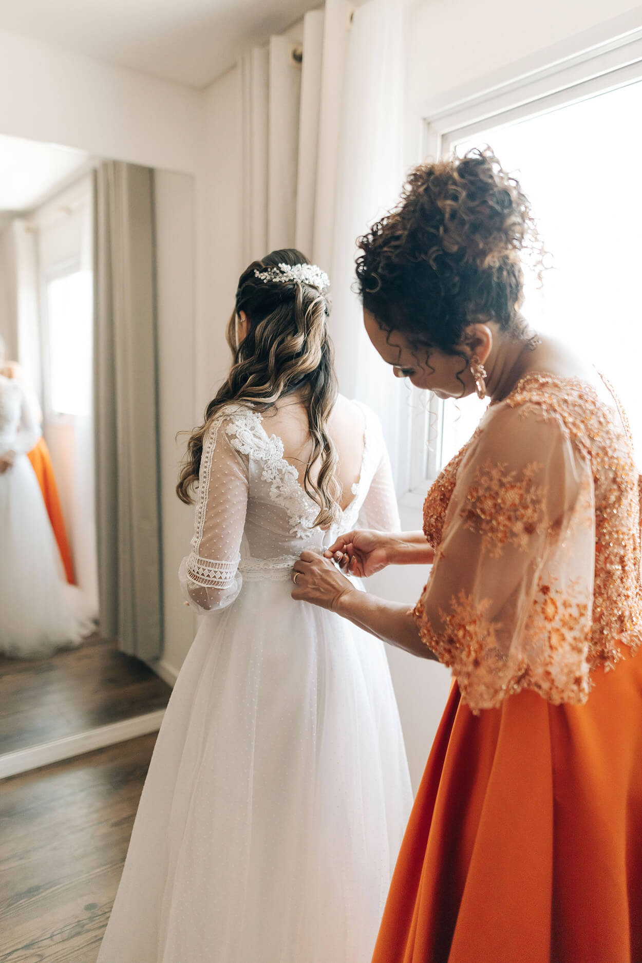Mulher com vestido laranja fechando o vestido da noiva no quarto