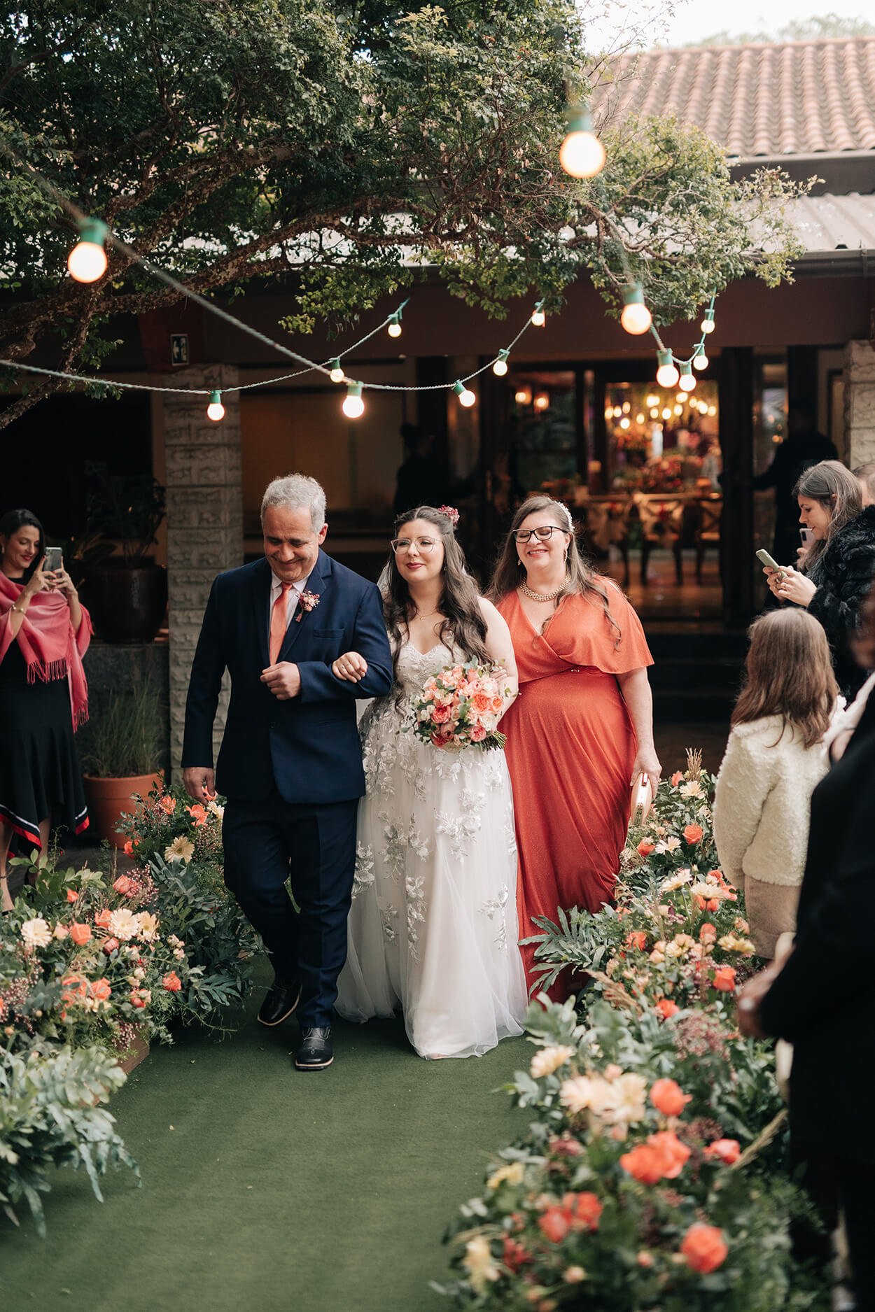 Noiva entrando com o seu pai de terno azul e sua mãe com vestido laranja