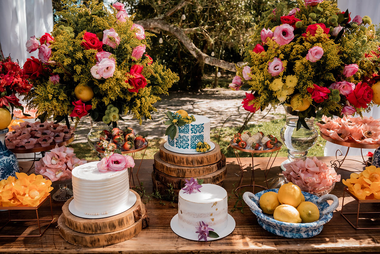 Mesa com três bolos de casamento branco com flores coloridas e arranjos florais com rosas vermelhas, rosas e flores amarelas