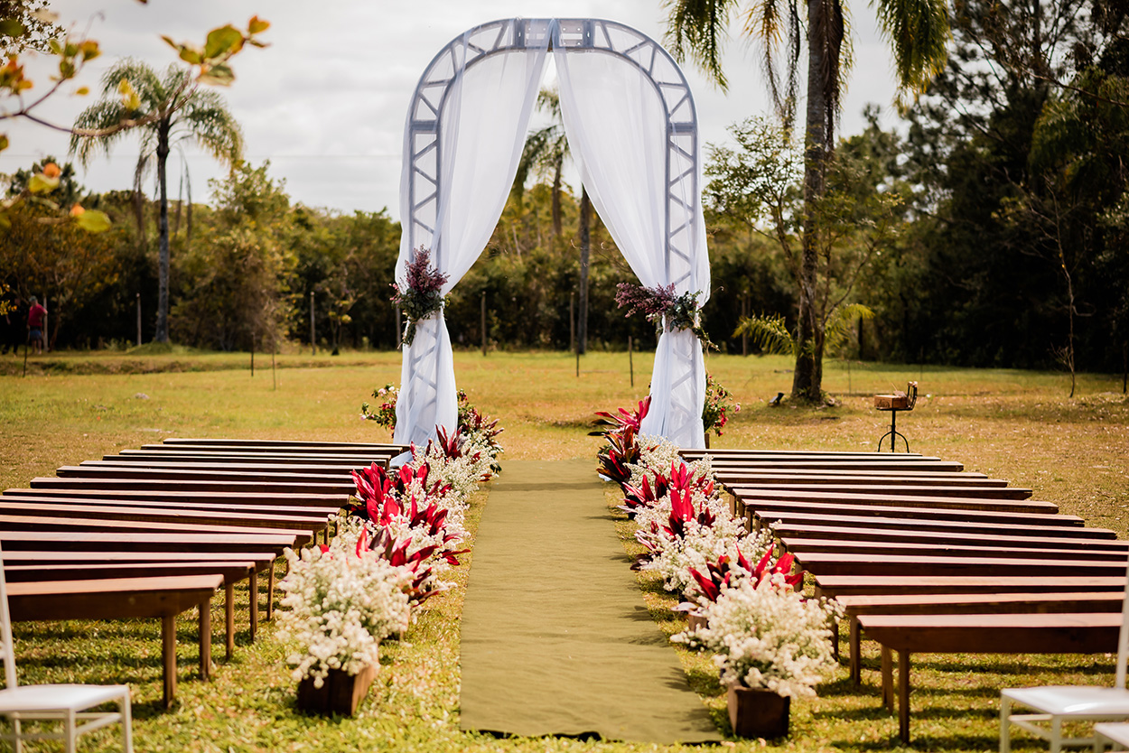 arco com cortinas brancas leves presas com arranjos e flores lilás e bancos de madeira no cmapo