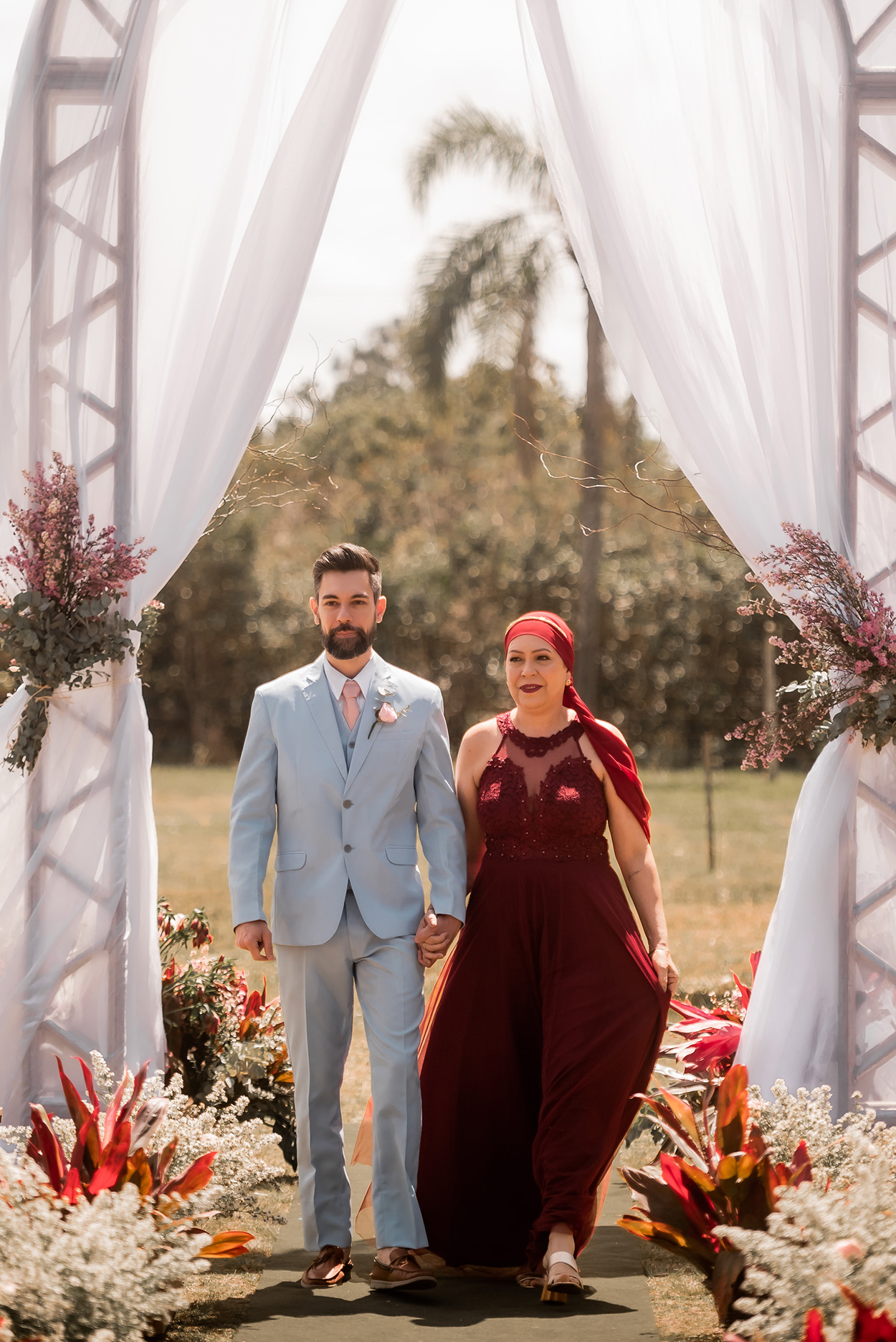 Entrada do noivo de terno azul com a mãe com vestido vermelho escuro leço da mesma cor na cabeça