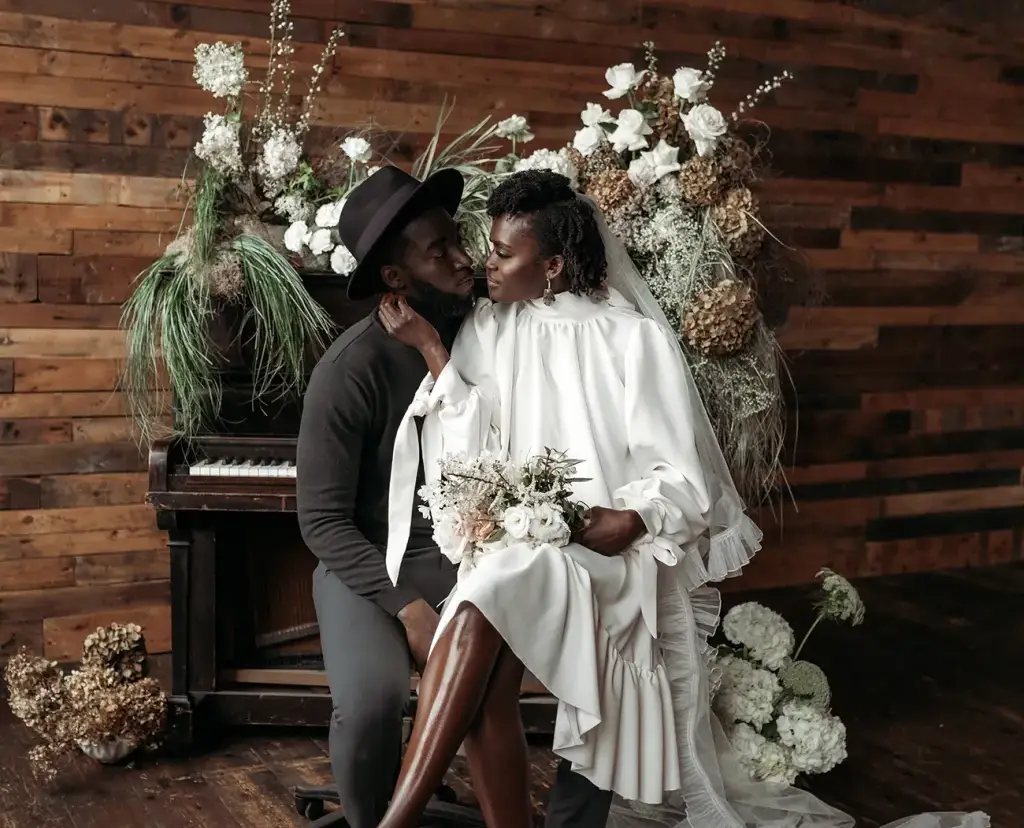 noiva negra com vestido curto e véu sentada no colo do noivo que veste roupa preta e chapéu em frente a um piano com flores