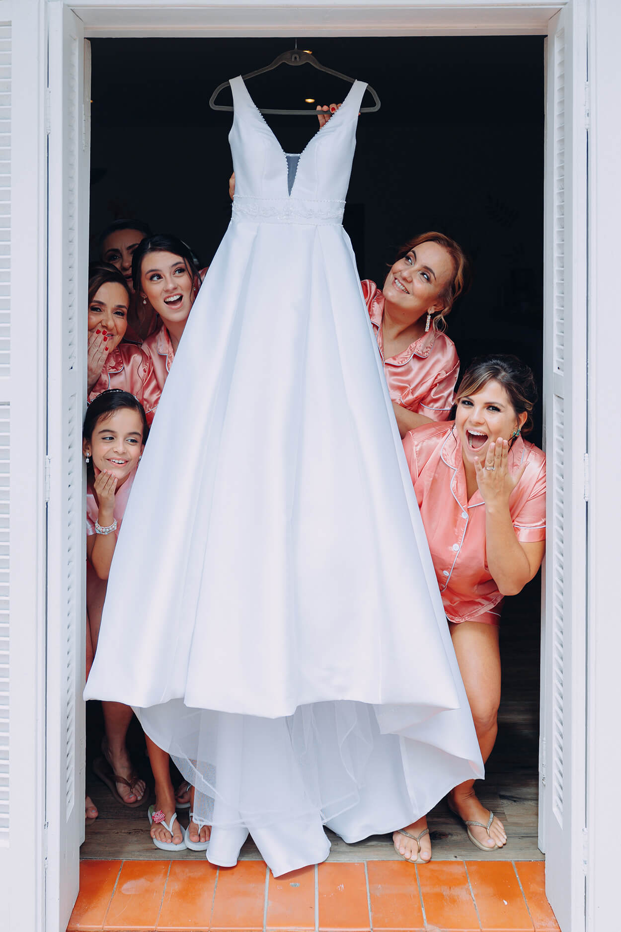 Madrinhas com robe cor de rosa atrás do vestidod e noiva pendurado na porta