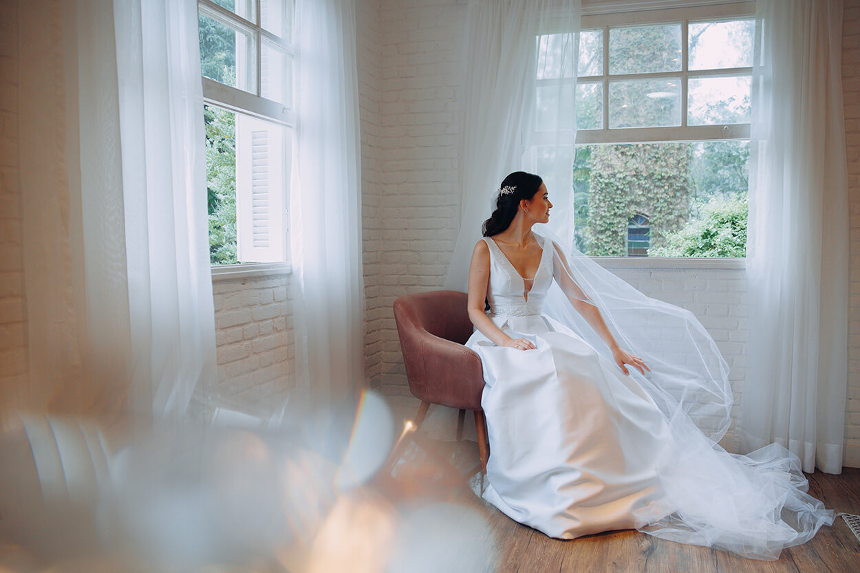Mulher com vestido de noiva clássico sentada em poltrona marrom dentro do quarto