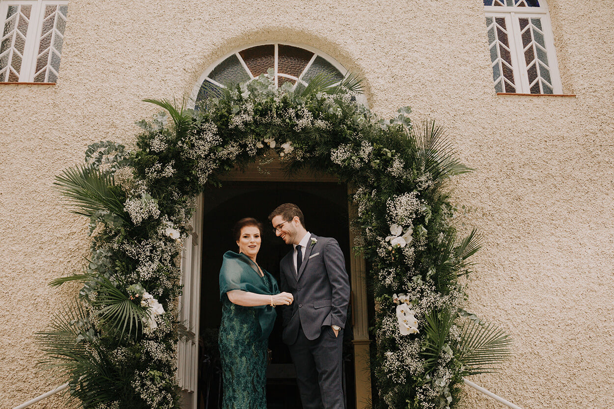 Noivo com terno cinza e mulher com vestido verde escuro an porta da capela