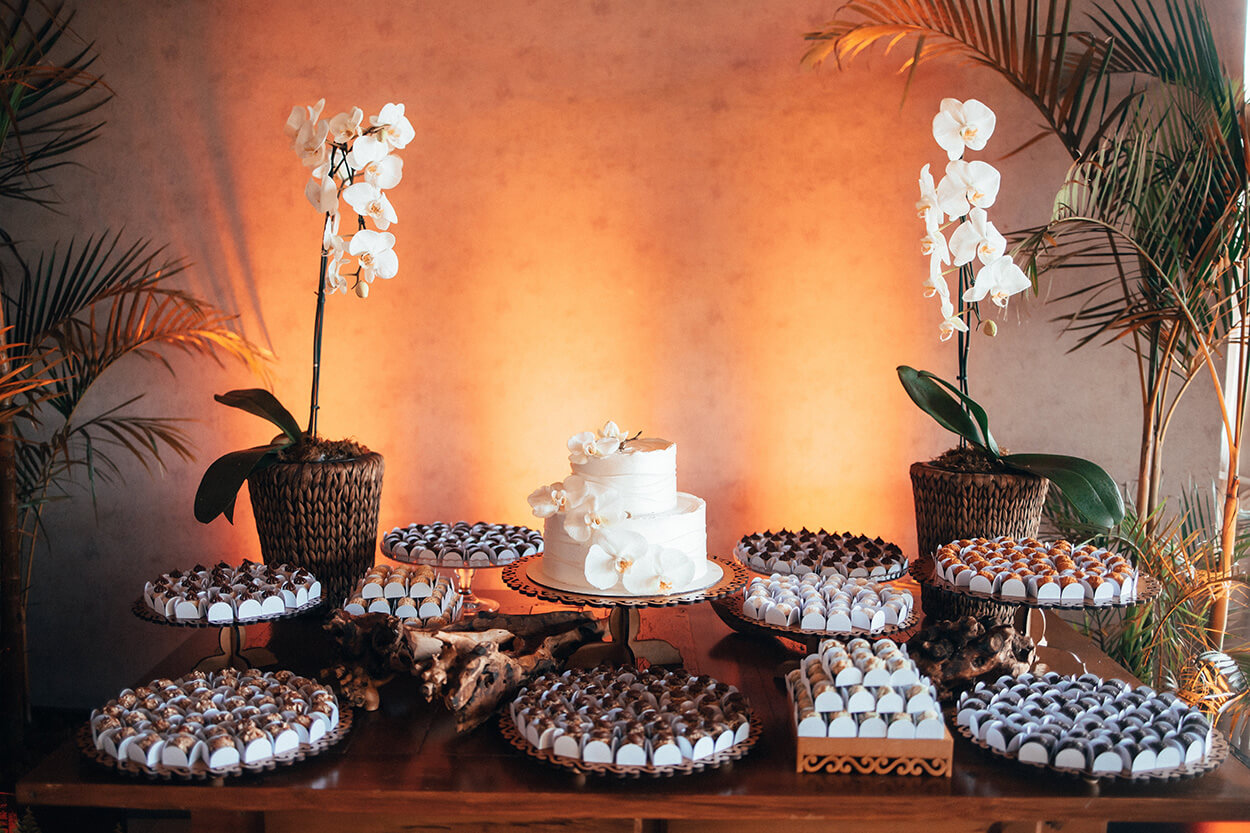 Mesa de madeira com bolo de casamento branco com orquídeas e travessas com doces