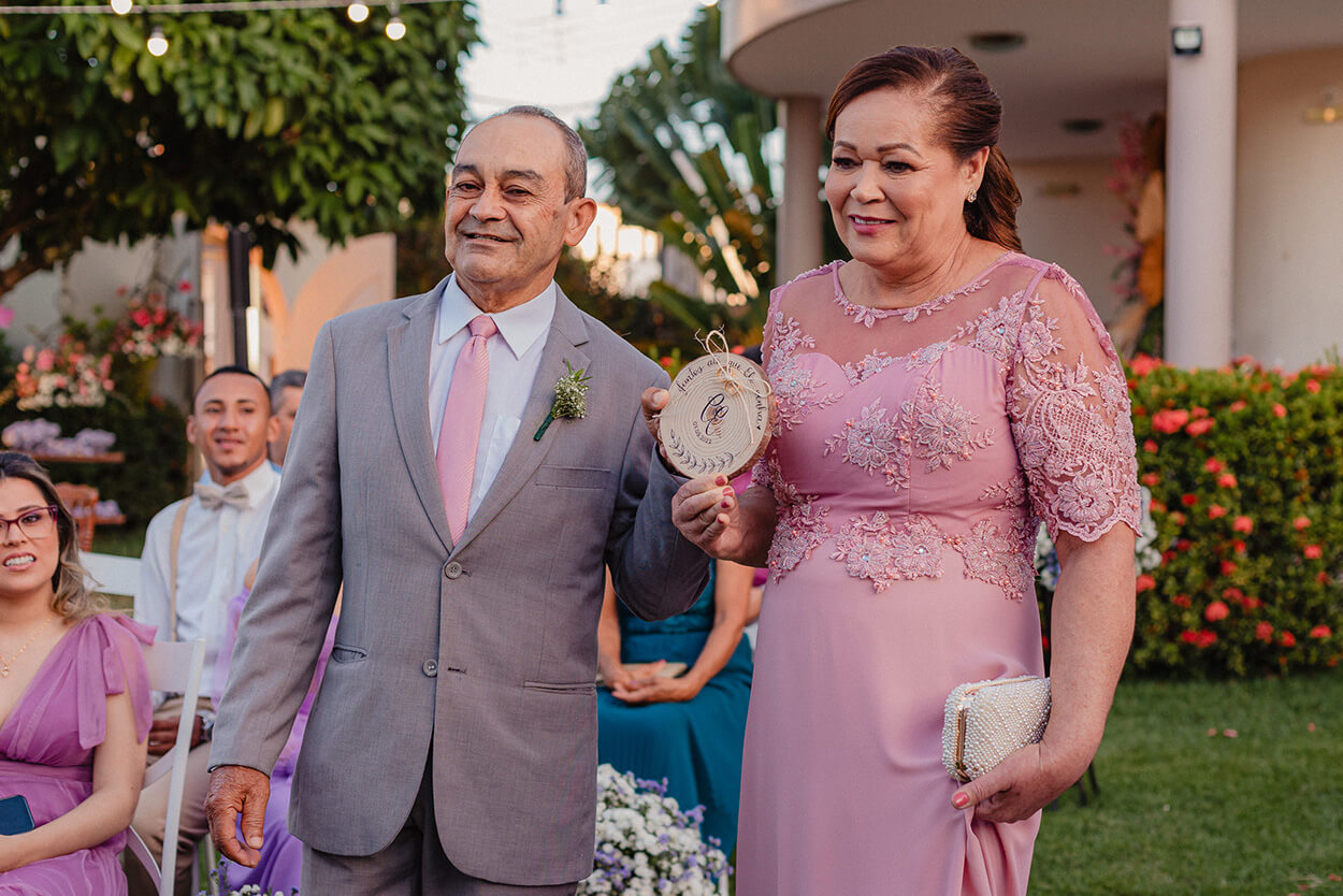 Pai do noivo com terno cinza e gravata rosa e mãe do noivo com vestido rosa com mangas
