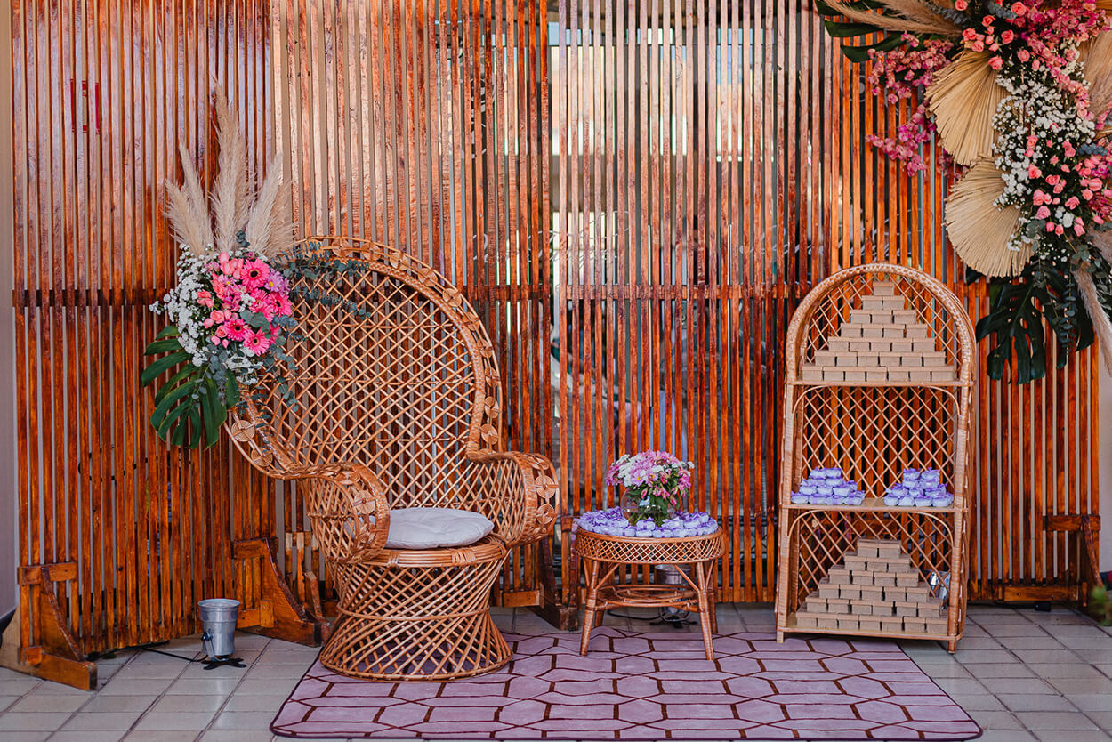 Cadeira pavão com capim dos pampas e flores rosas ao lado de mesinha e estante com lembrancinhas