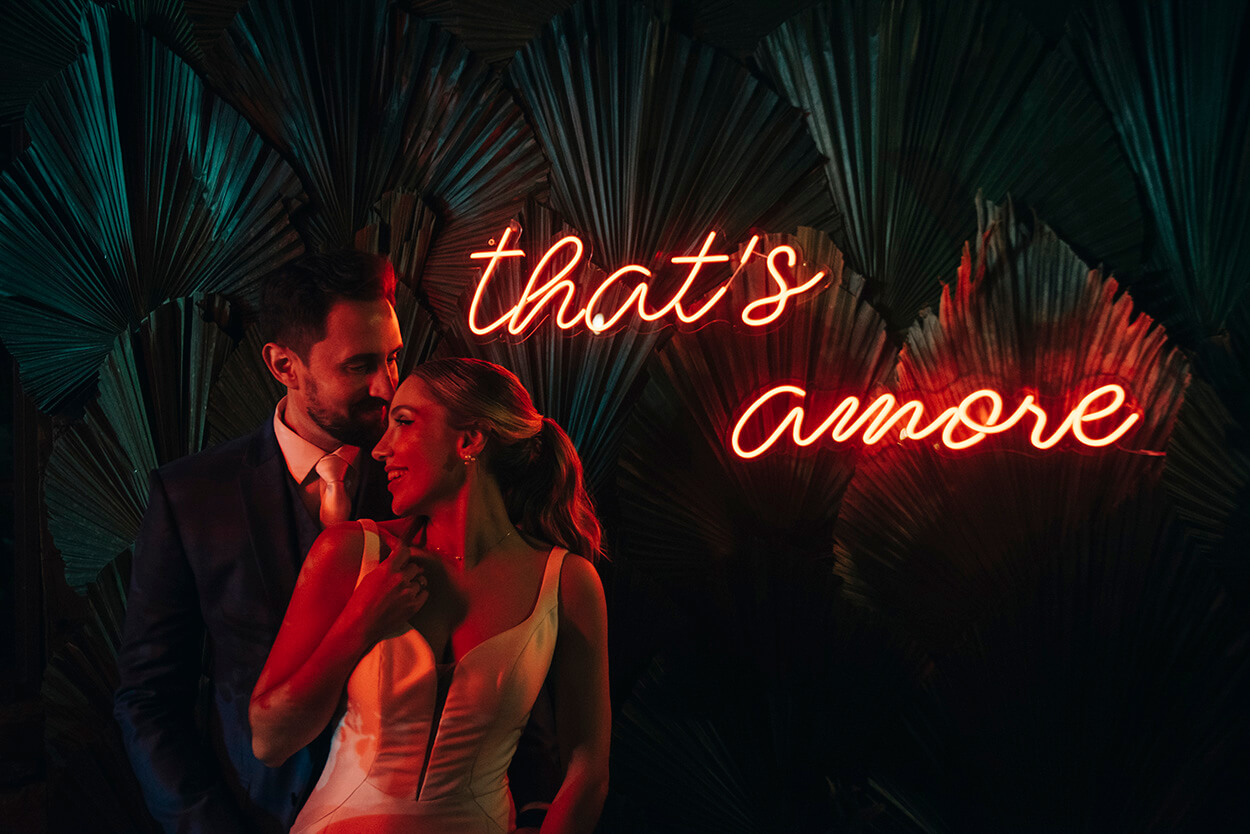 Noivos ao lado de painel neon vermelho escrito that's amore