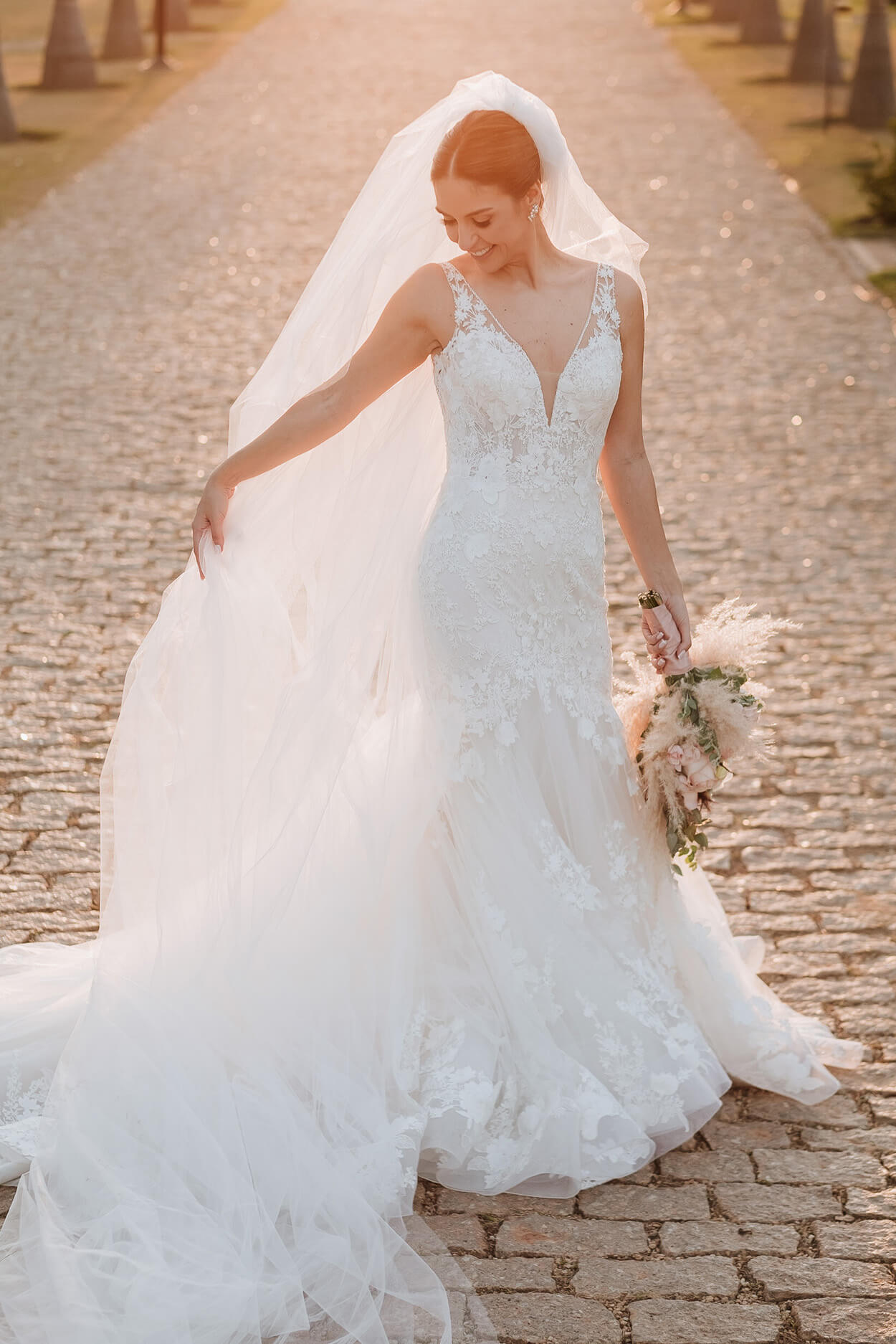 Mulher com vestido de noiva com cauda e véu longo segurando buquê ao ar livre