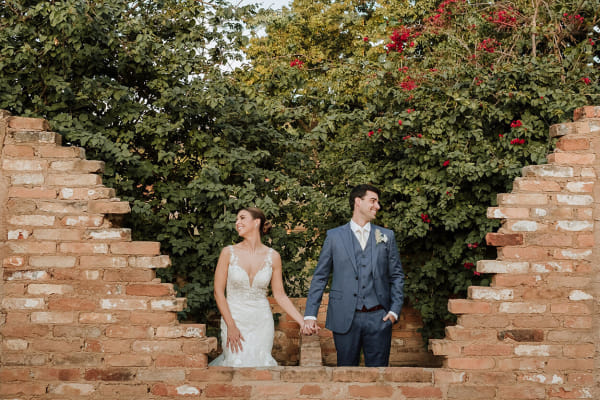 capa post: Noivos de mãos dadas olhando para os lados atrás de muro em ruínas ao ar livre