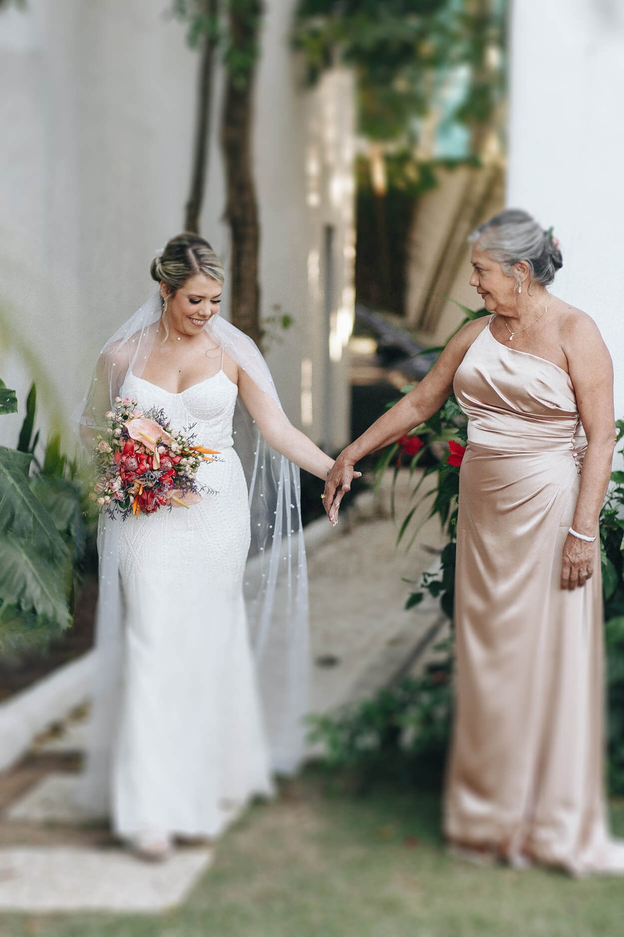 Noiva com buquê colorido de mãos dadas com sua mãe com vestido rosê cintilante