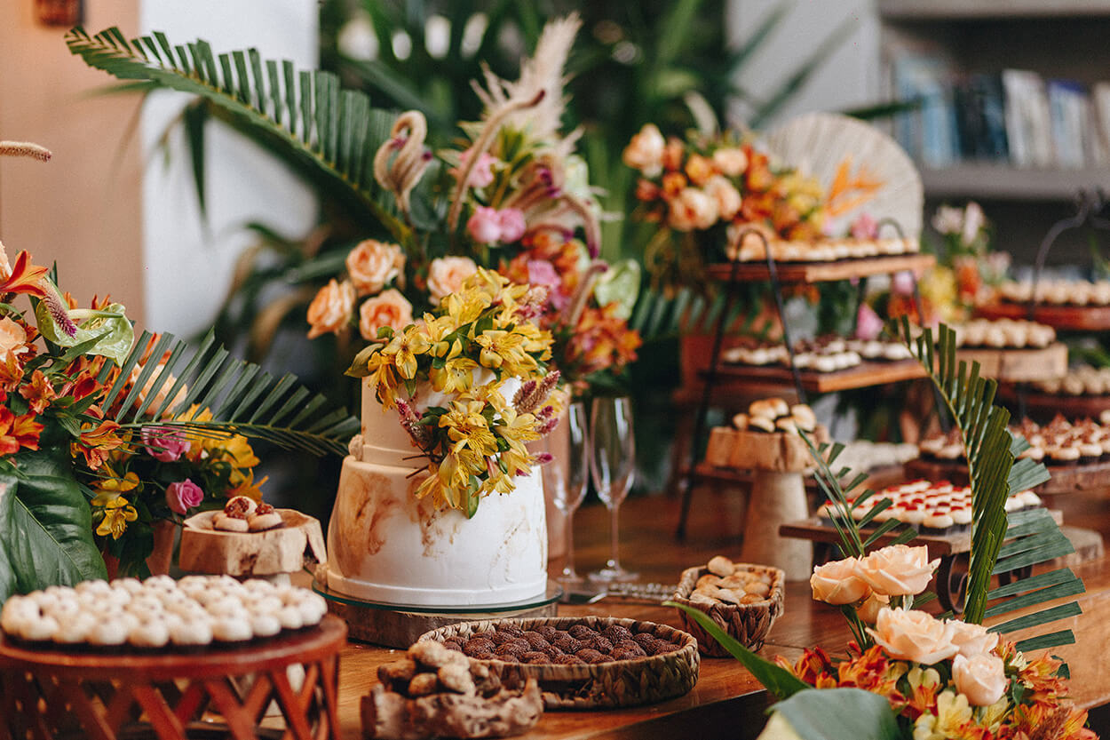 Mesa de madeira com bolo de casamento branco com flores amarelas e bandejas com docinhos