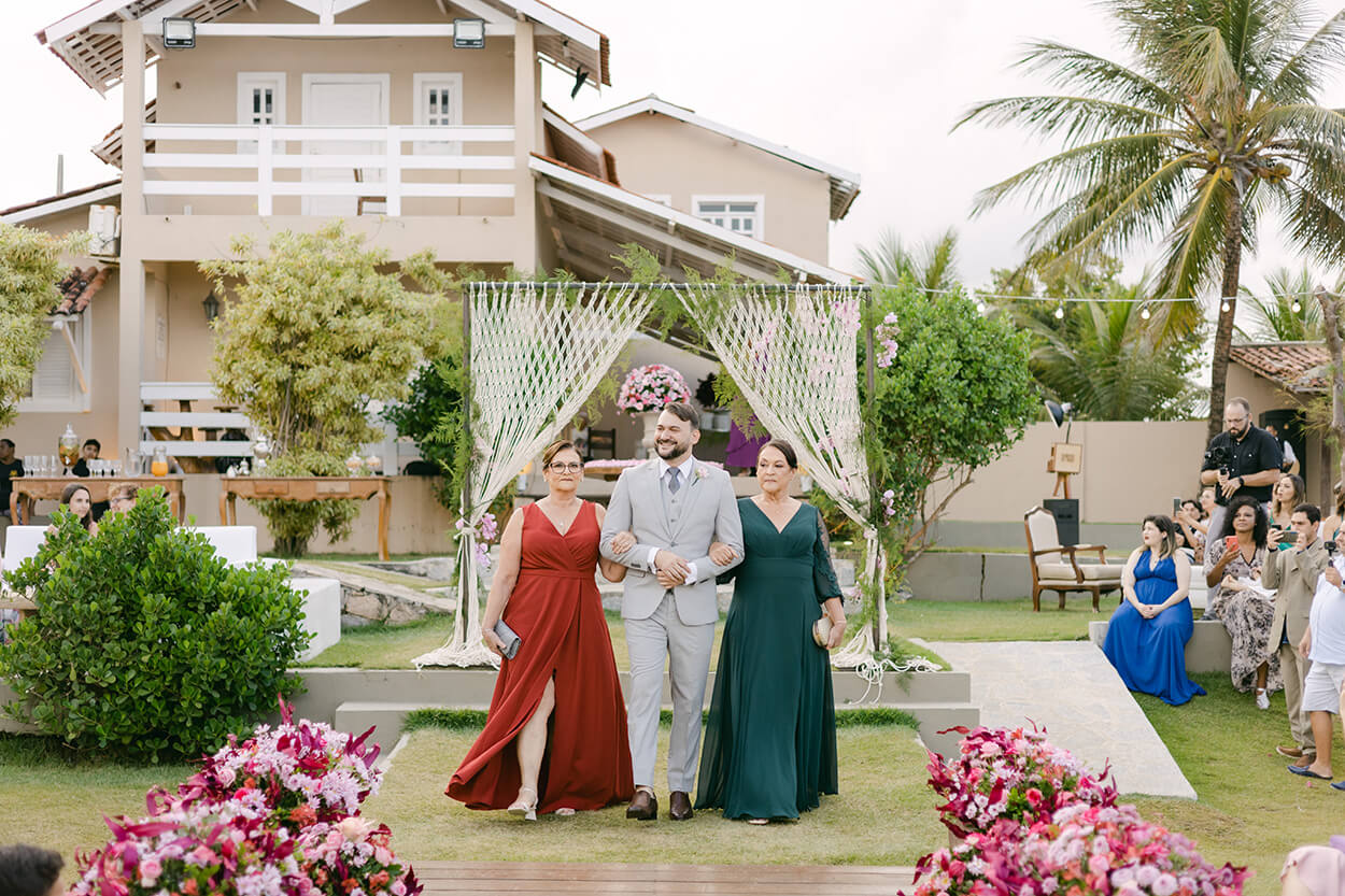Noivo entrando no casamento com mulher de vestido laranja e outra com vestido verde esmeralda