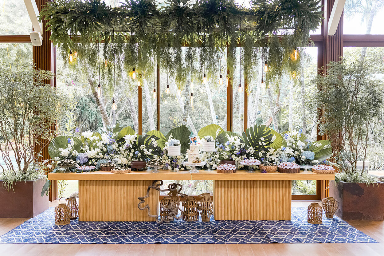 Mesa do bolo de madeira com trio de bolos de casamento branco, doces decorada com palmeiras leque, costela-de-adão e hortênsias azuis