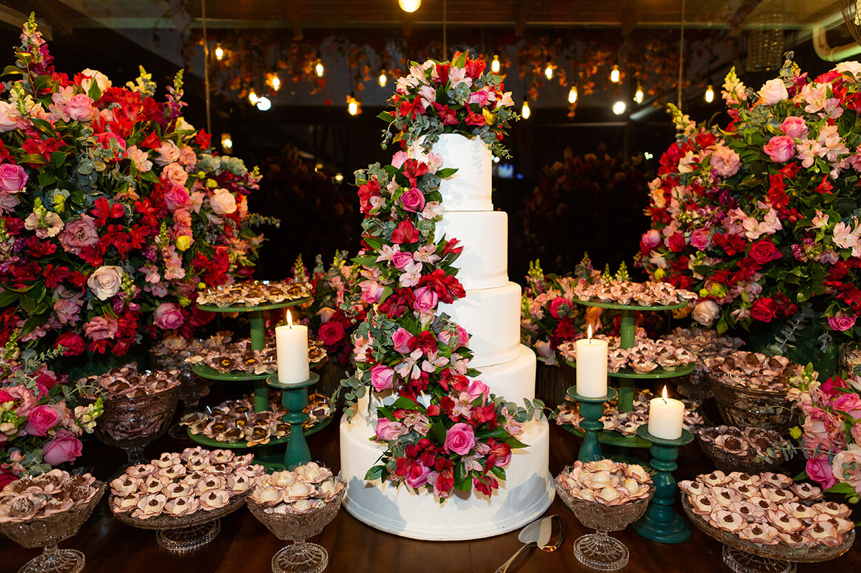 Mesa de madeira com bolo de casamento branco com cinco andares e topo decorado com rosas e outras flores