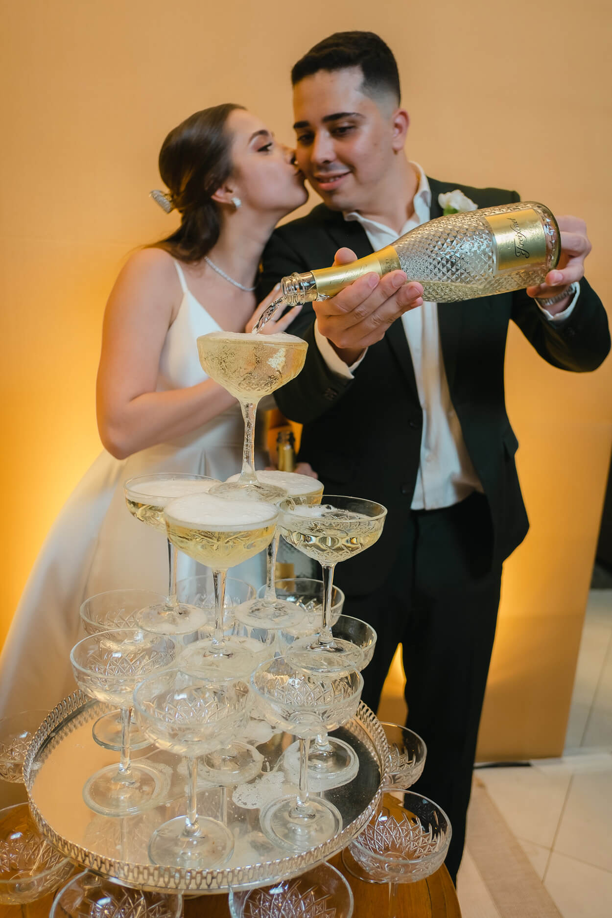 Noiva beijando o rosto do noivo enquanto ele despeja bebida na torre de taças