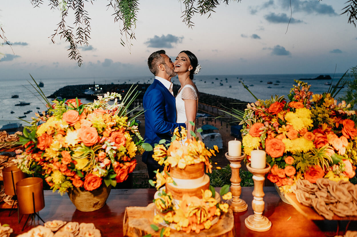 Noivo abraçandoe beijando o rosto da noiva atrás da mesa com bolo de casamento