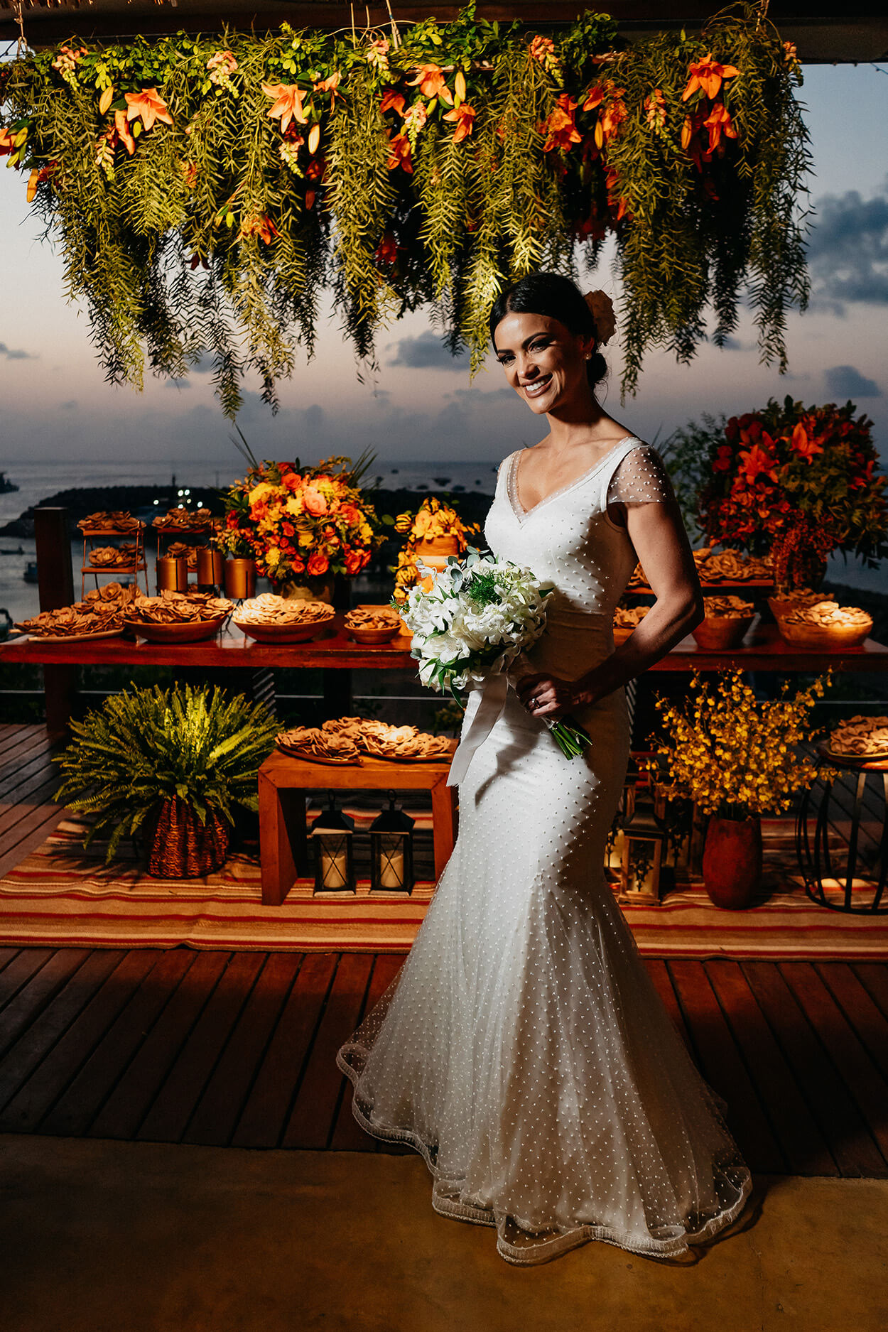 Noiva sorrindo e segurando buquê com flores brancas na frente da mesa de bolo de casamento