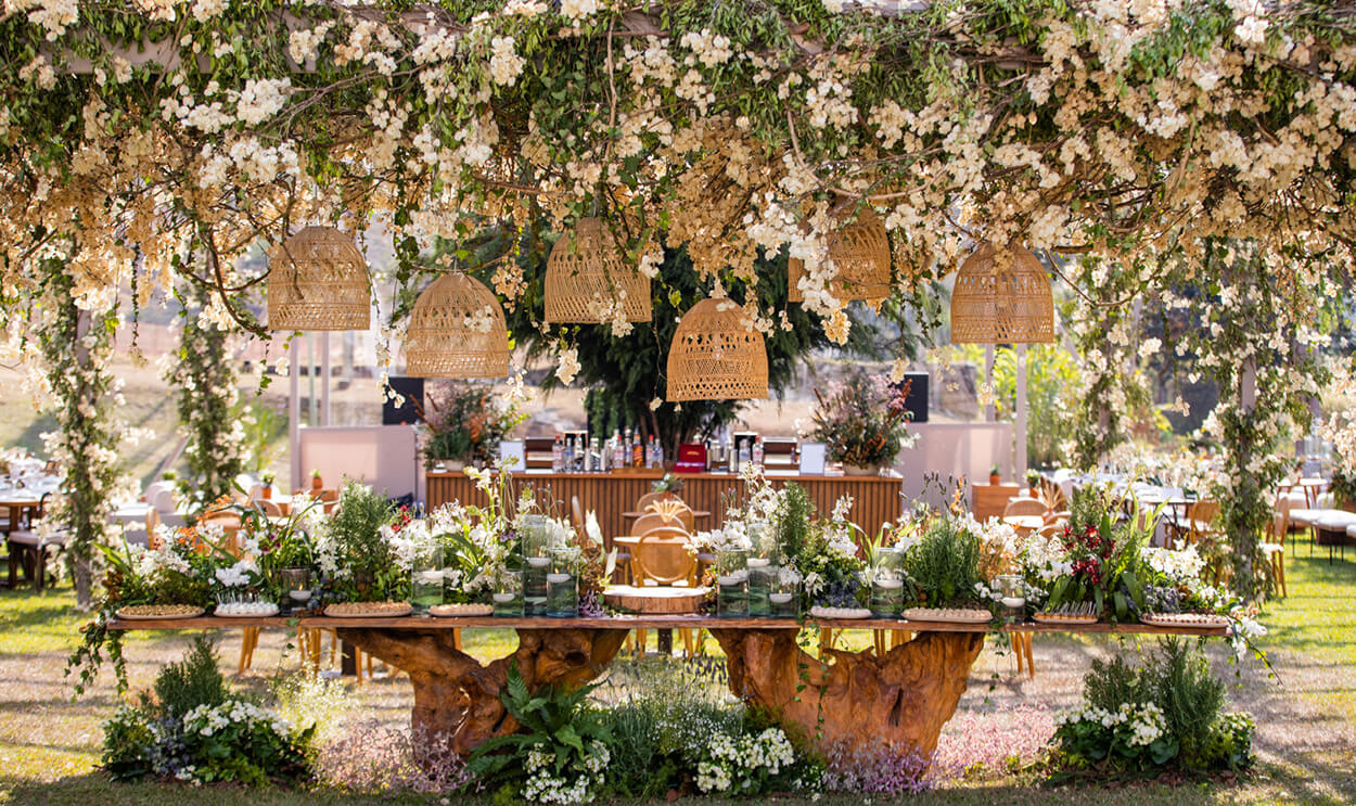 Mesa rústica com doces de casamento, vidros com água e velas, e luminárias do tipo cesta artesanala e flores brancas pendentes