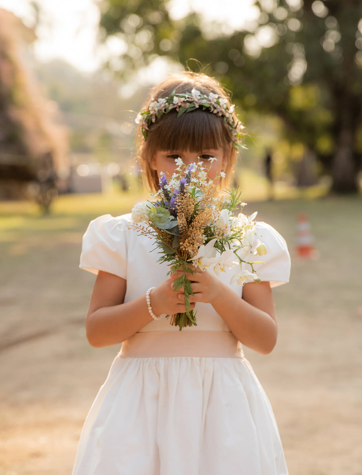 Daminha com vestdio branco com fita rosa escondendo o rosto com mini buquê brancoe lilás