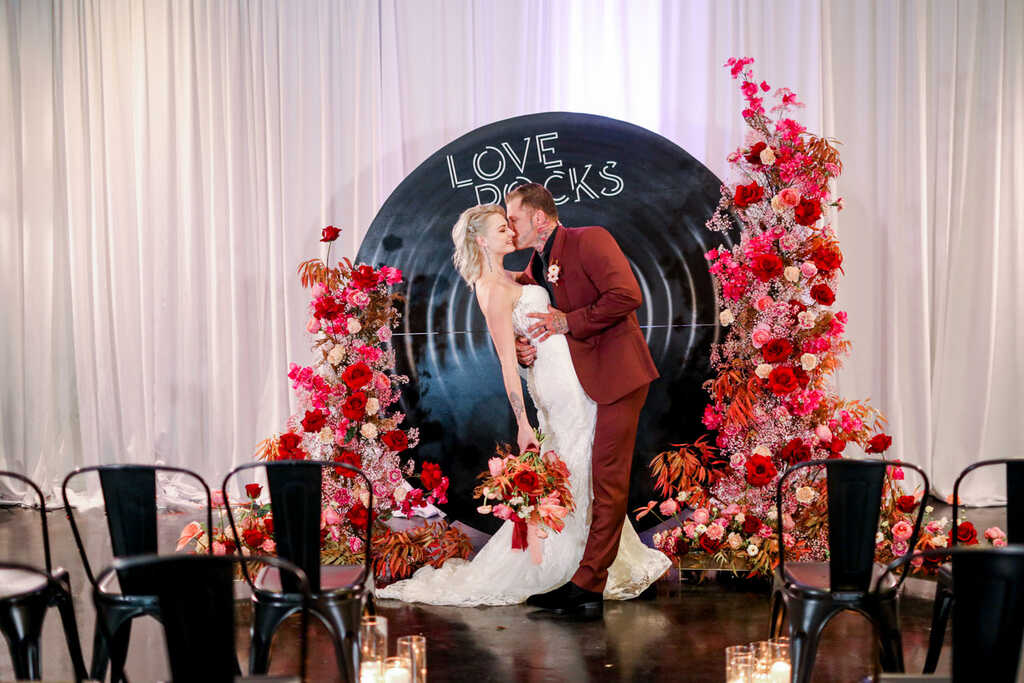 noivos se beijando em frente ao painel em formato de disco de vinil com arranjos florais