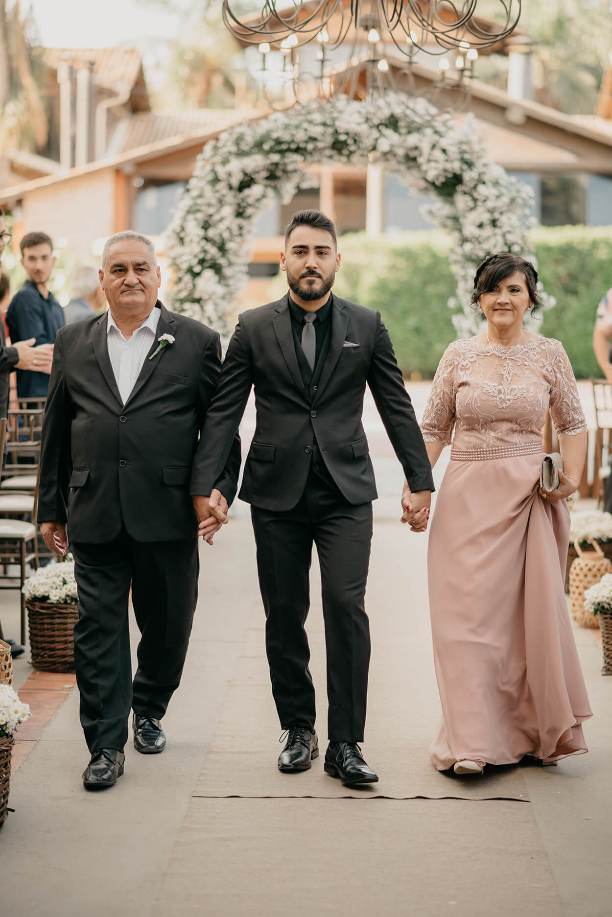 Noivo caminhando até o altar de mãos dadas com o sue pai de terno preto e mãe com vestido longo rosê