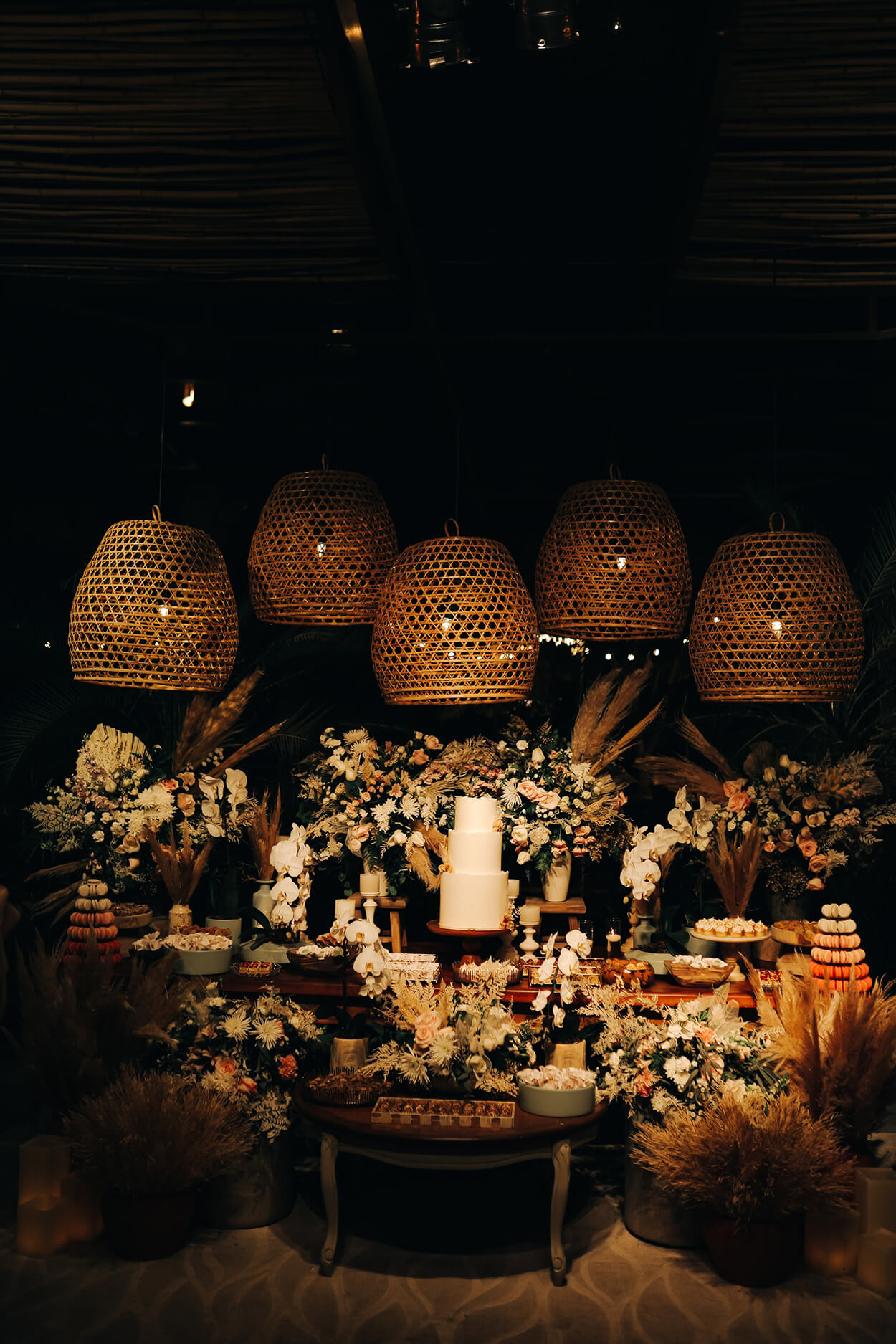 Mesa com bolo de casamento branco com arranjos com rosas e grandes luminárias redondas