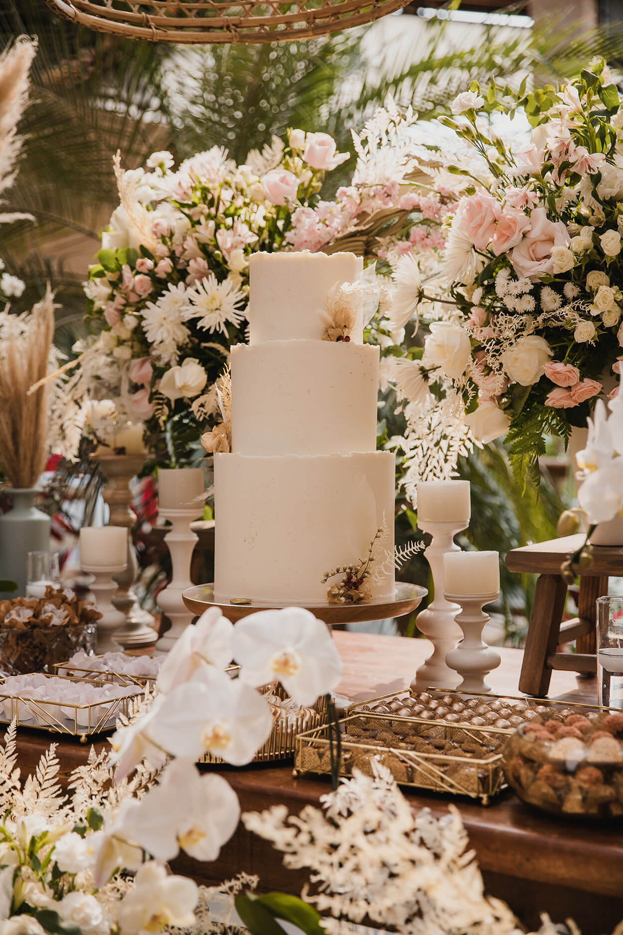 Mesa de madeira com orquídeas brancas arranjos com rosas e bolo de casamento branco redondo com três andares