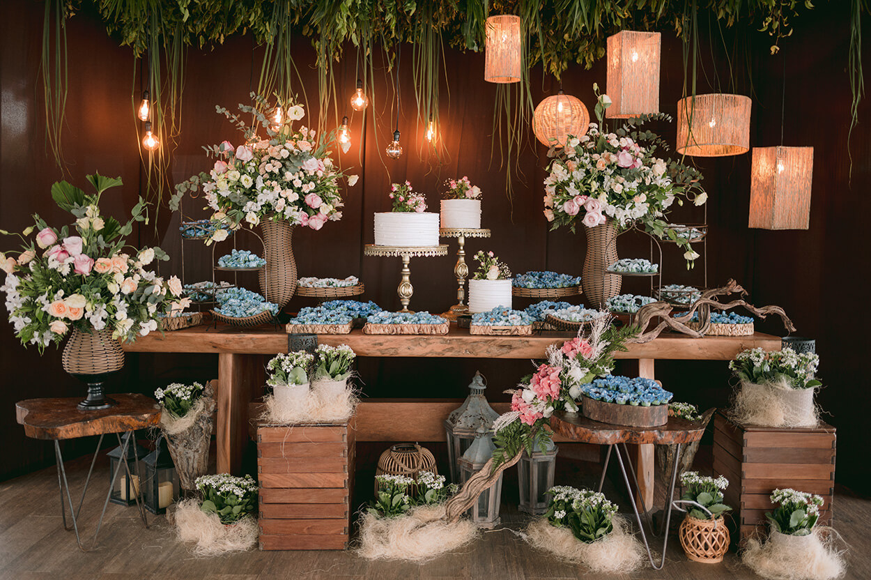 Mesa de madeira com bandejas com doces de casamento com forminhas azuis serenity arranjos de flores brancas e rosas e trio de bolos brancos