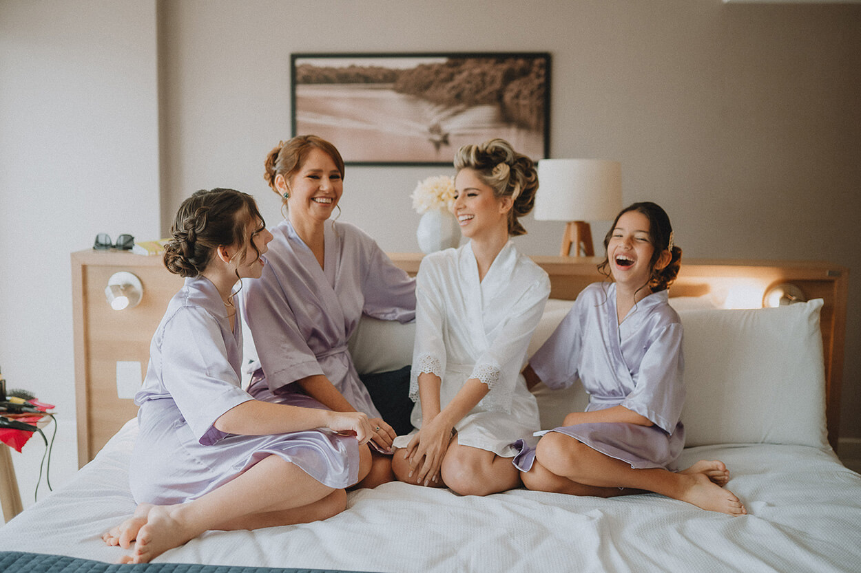 Noiva sentada na cama com robe branco ao lado de três moças com robe lilás
