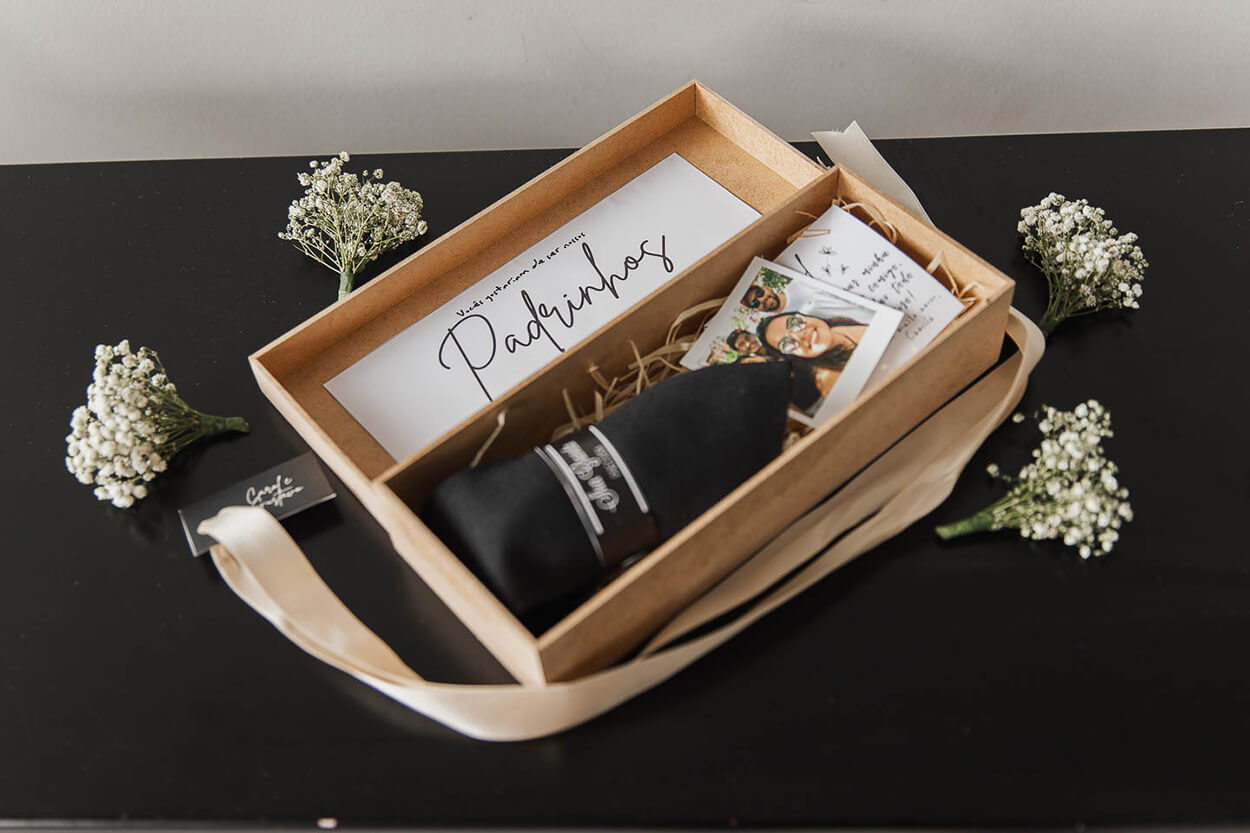 Caixa de madeira de presente para padrinhos cartinha e fotos em cima de mesa preta fosca e flroes brancas ao lado