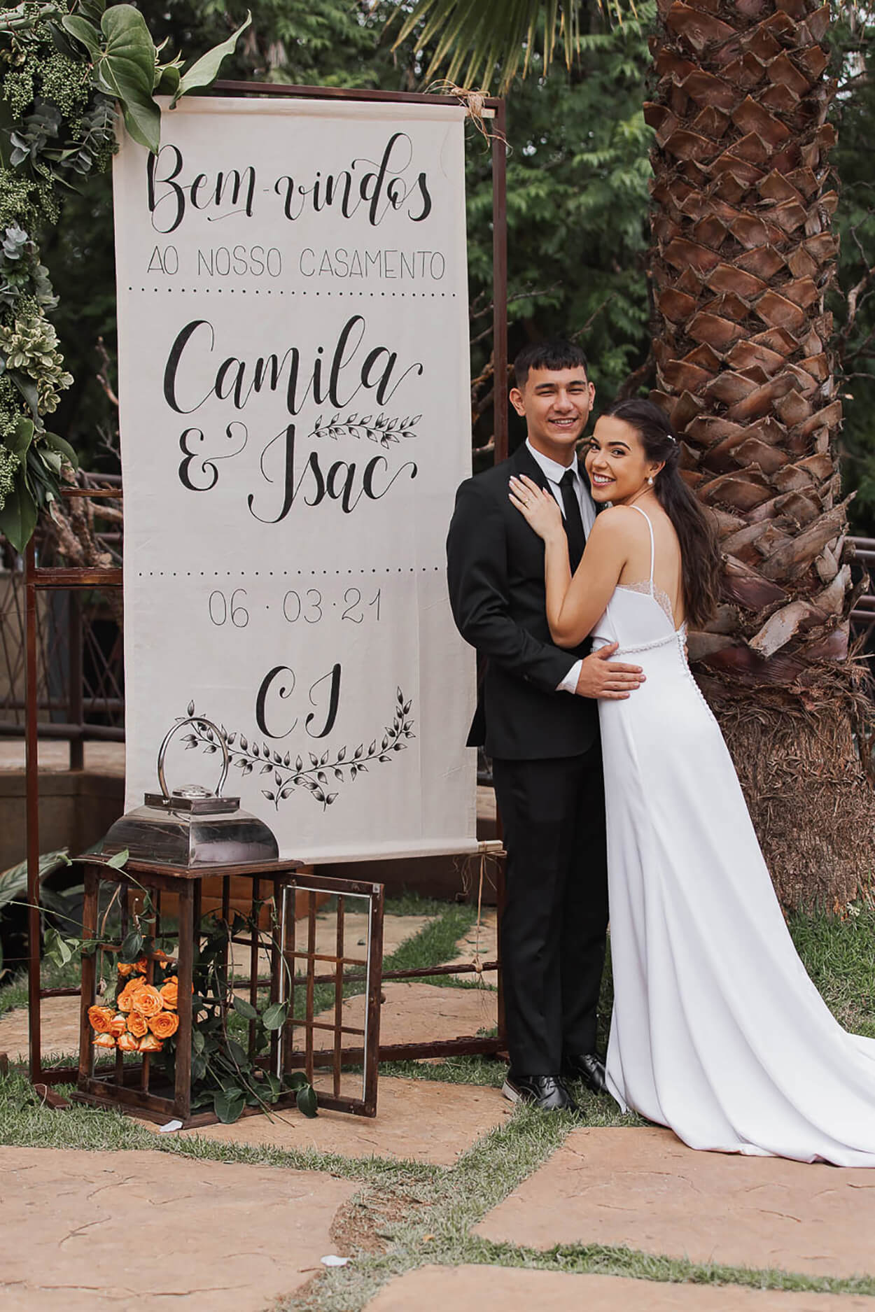 Noivos abraçados ao lado de placa de bem-vindos escrito Camila e Isac em letras cursivas no jardim