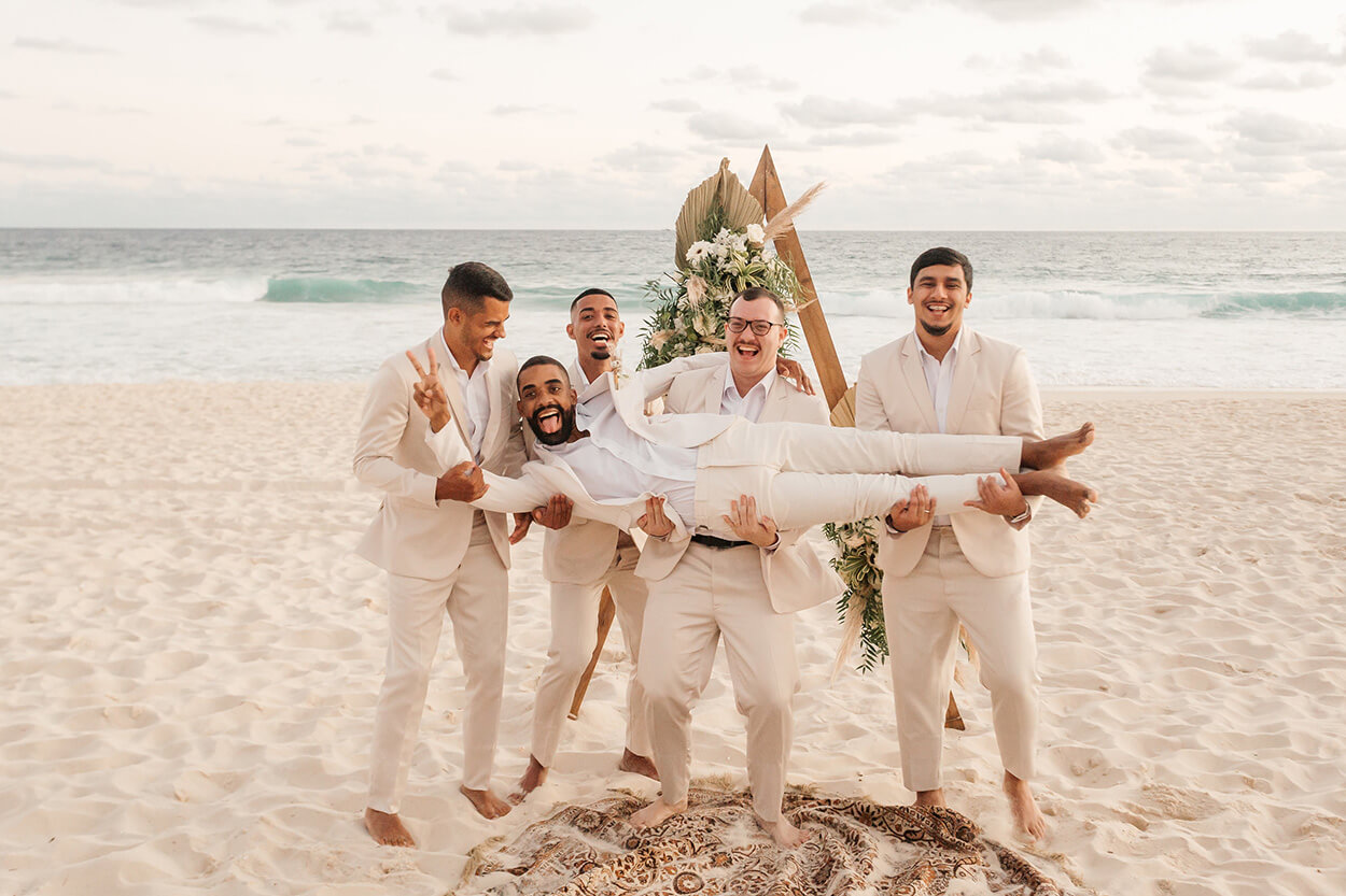 Padrinhos com terno e calça off white carregando o noivo no colo na praia