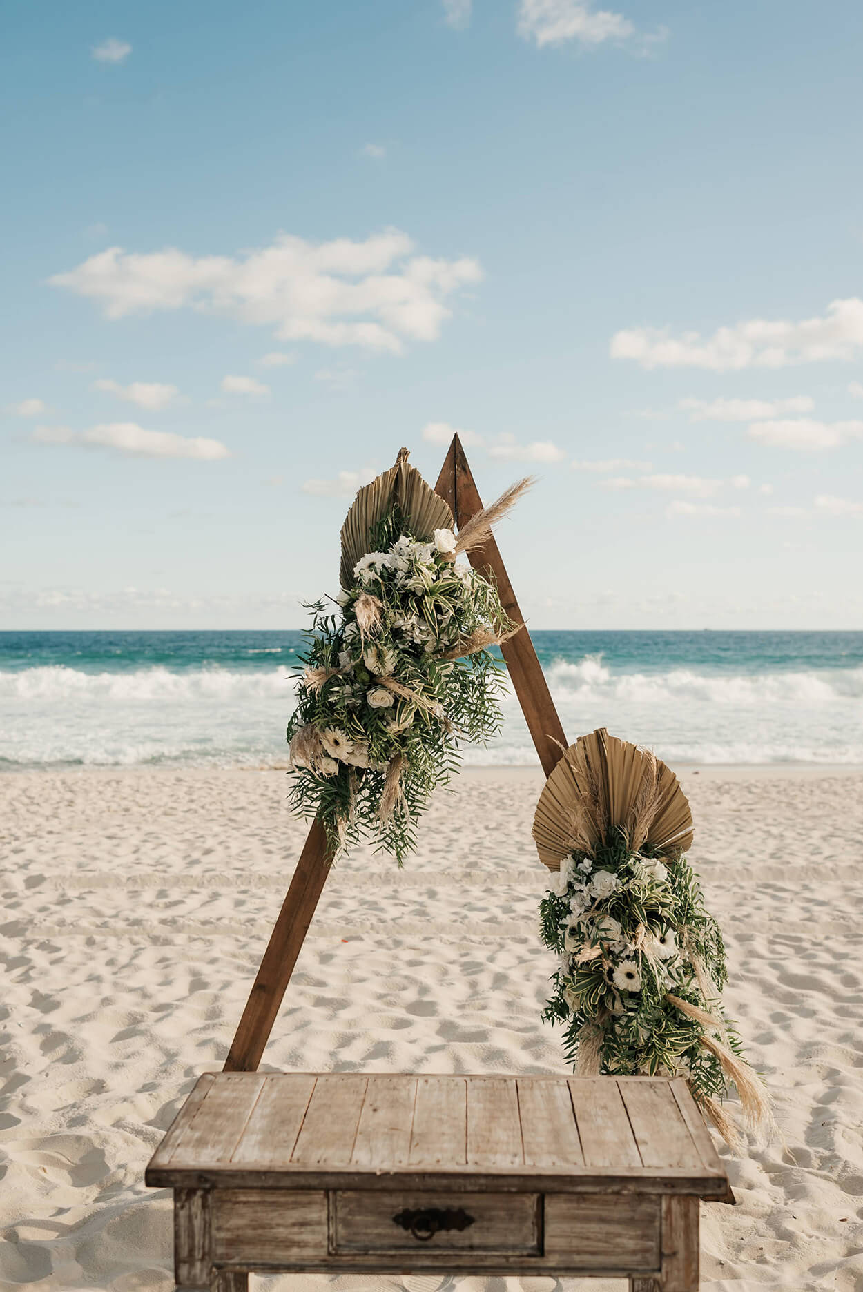 Altar triangular de madiera com arranjo de flores boho na areia da praia