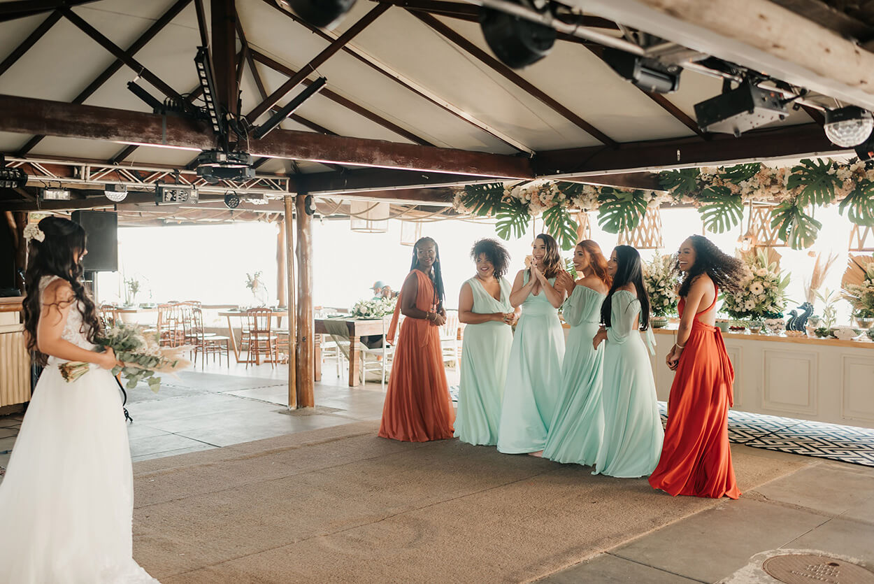 Quatro madrinhas com vestidos azuis claros e duas com vestido terracota olham para trás para ver a noiva