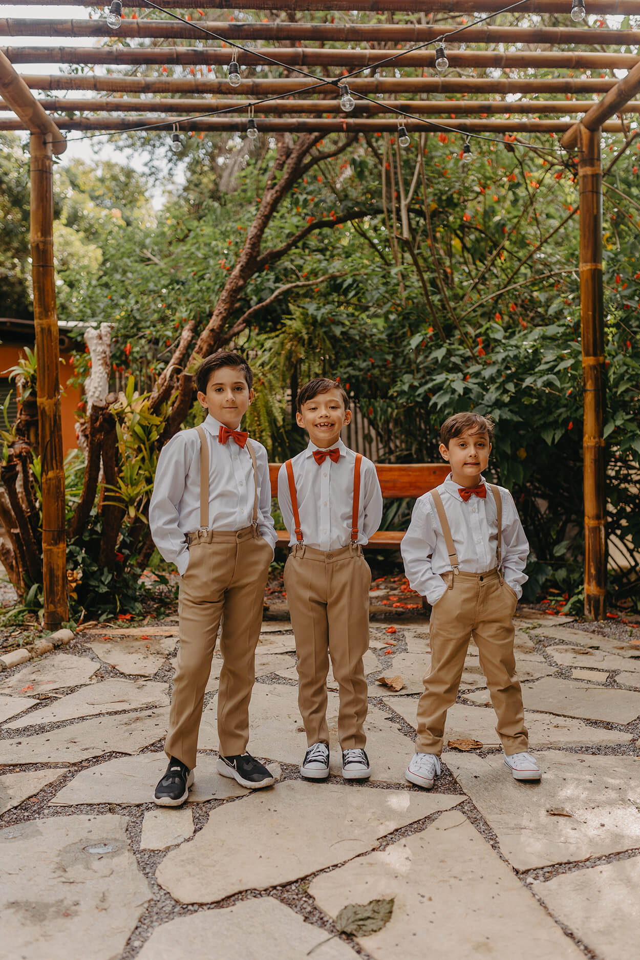 três meninos com calça marrom com camisa branca com suspensório e gravatas laranjas no jardim