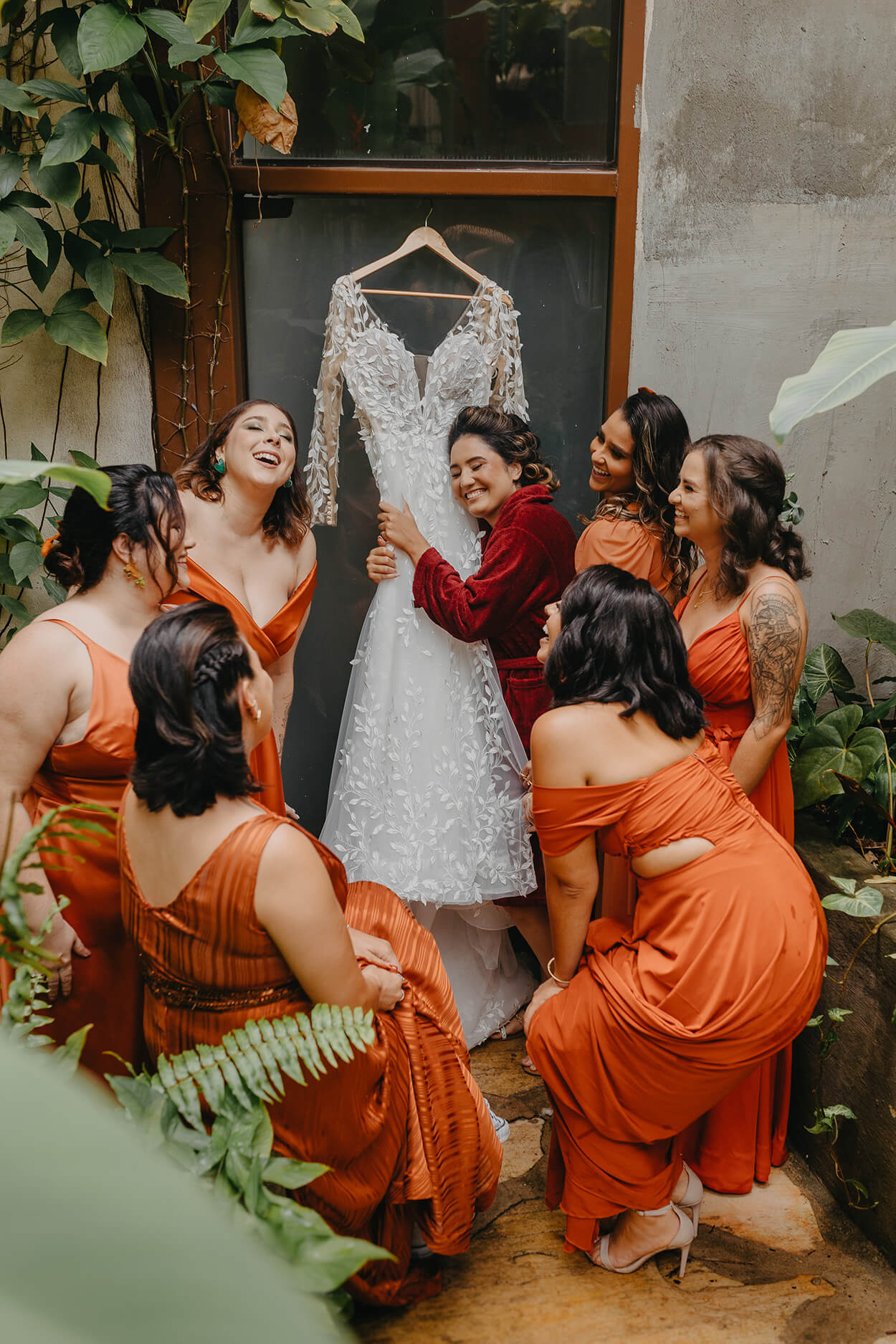Noiva com robe vermelho abraçando o vestido de noiva e madrinhas com vetsido laranja em volta