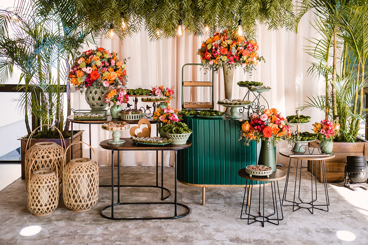 Salão com mesas modernas e arredondadas com bandejas com doces de casamento e vasos com flores vermelhas, rosas e laranjas