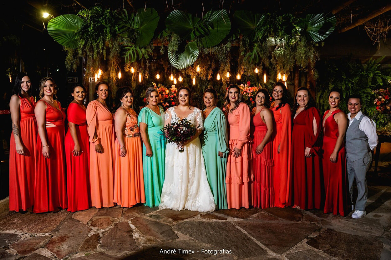 Madrinhas com vestidos longos vermelhos, laranjas e azuis ao lado da noiva 