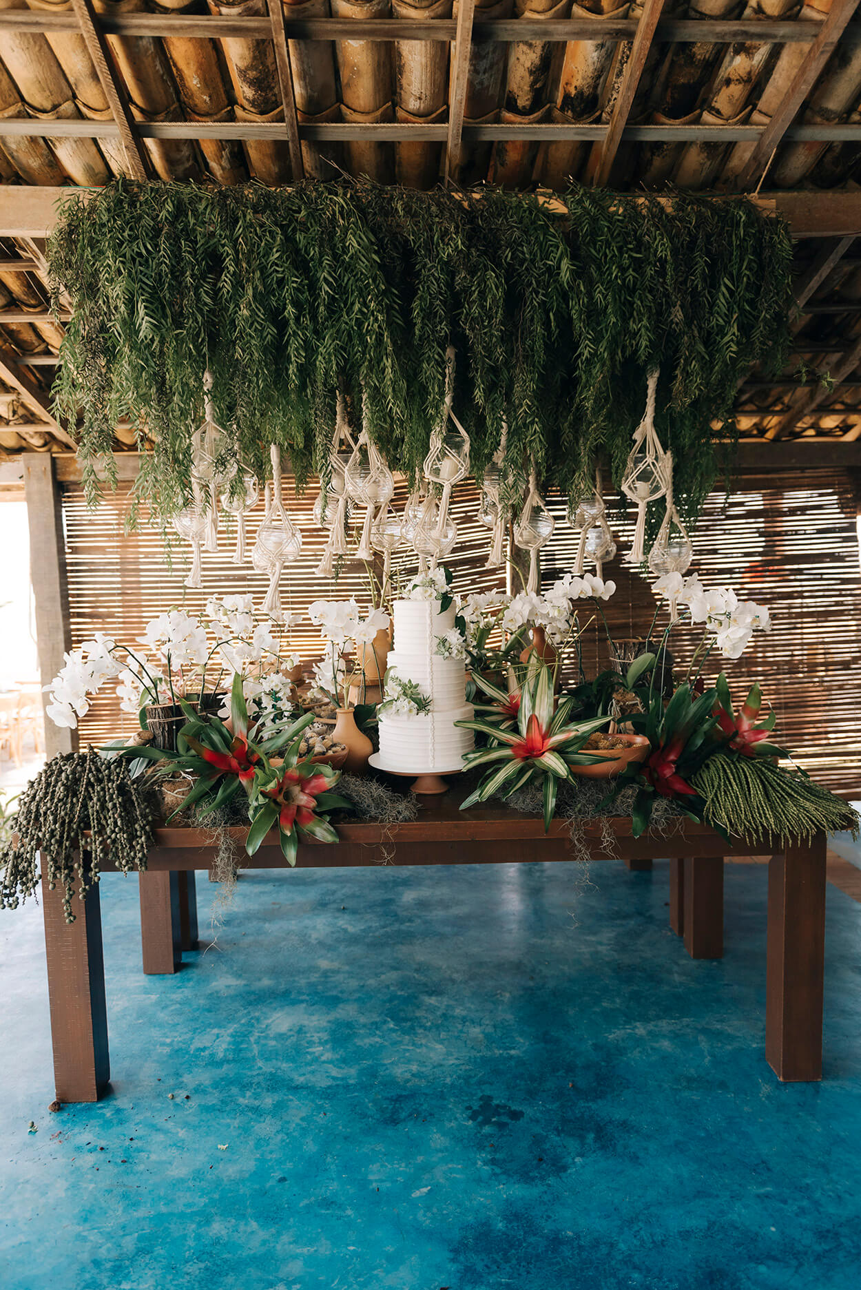 Salão com chão azul e mesa de madeira com orquídeas brancas folhagens e um bolo de casamento branco com três andares e flores