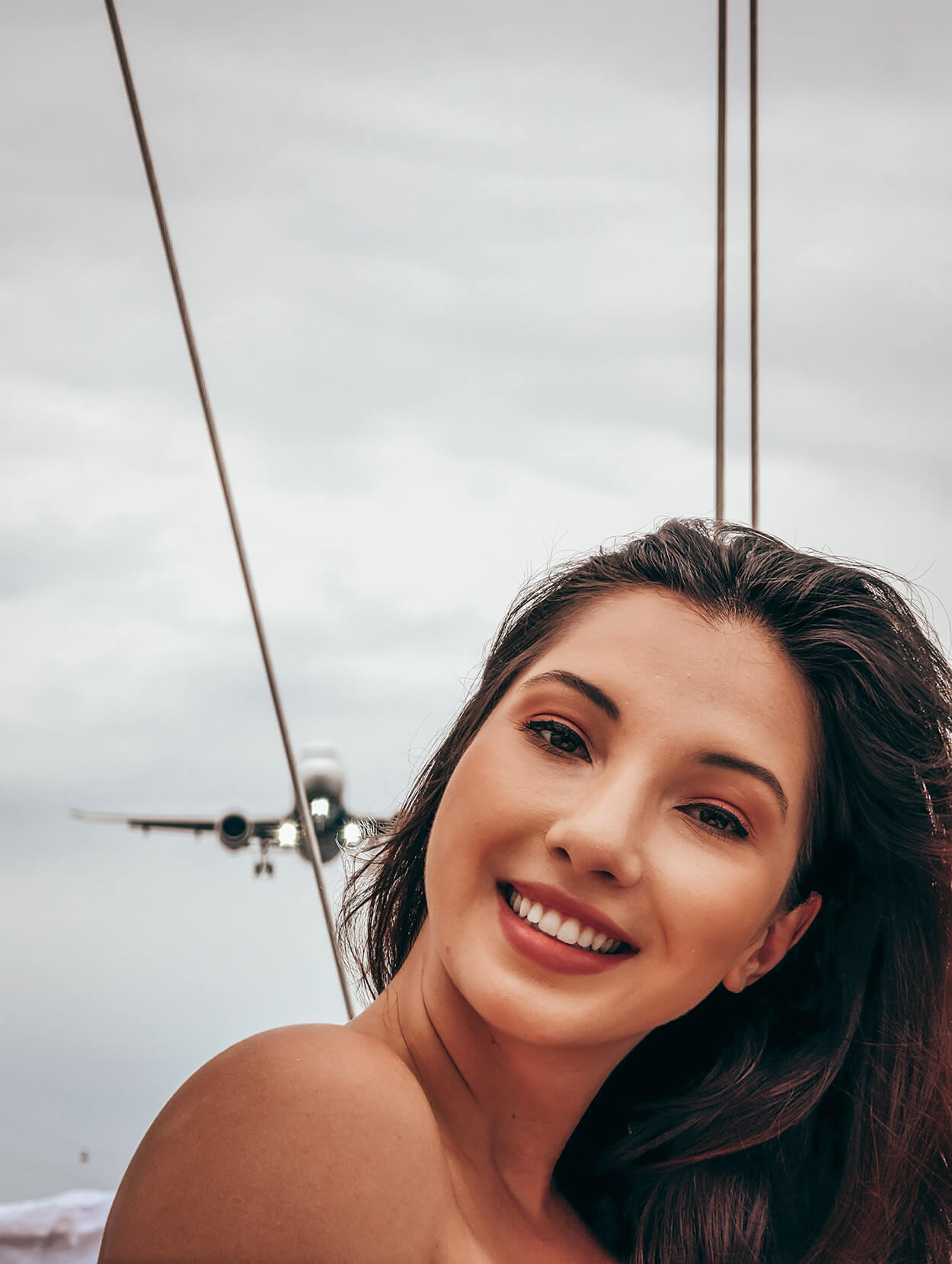 Mulher jovem dde cabelos castanhos sorrindo enquanto avião passa ao fundo