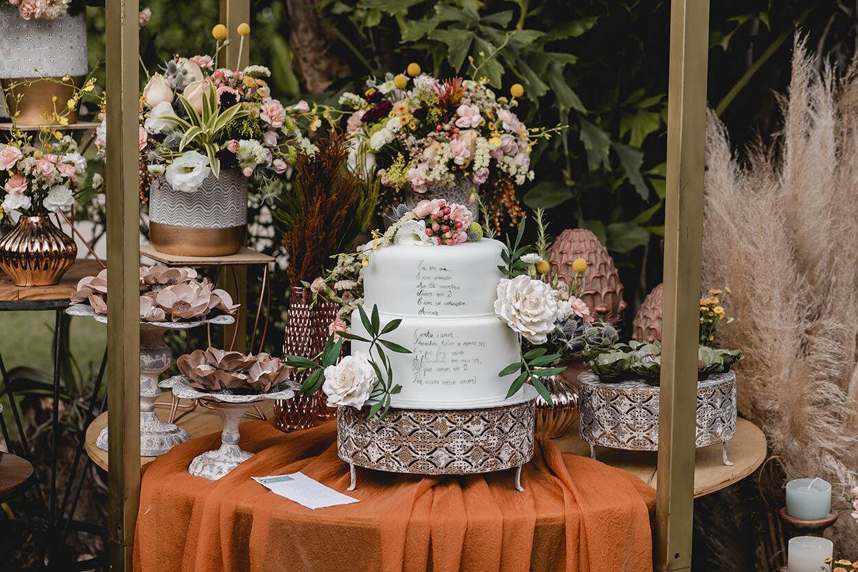 Mesa com tecido laranja e bolo de casamento de dois andares com poema escrito além de rosas e flores no topo
