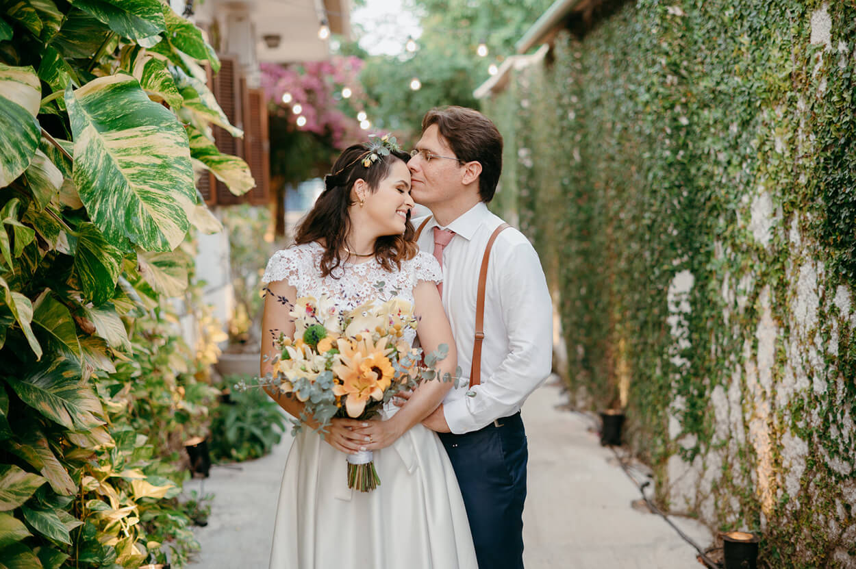 Noivo beijando a testa da noiva em um corredor cheio de plantas