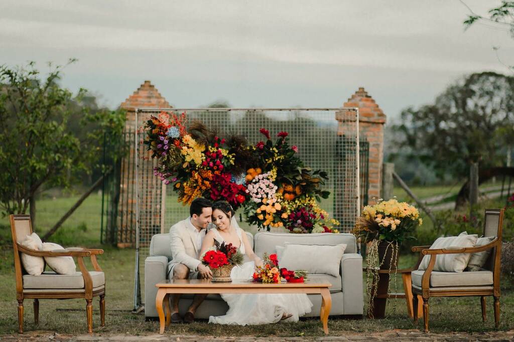 noivos sentados em sofá com painel de flores em decoração de casamento colorida