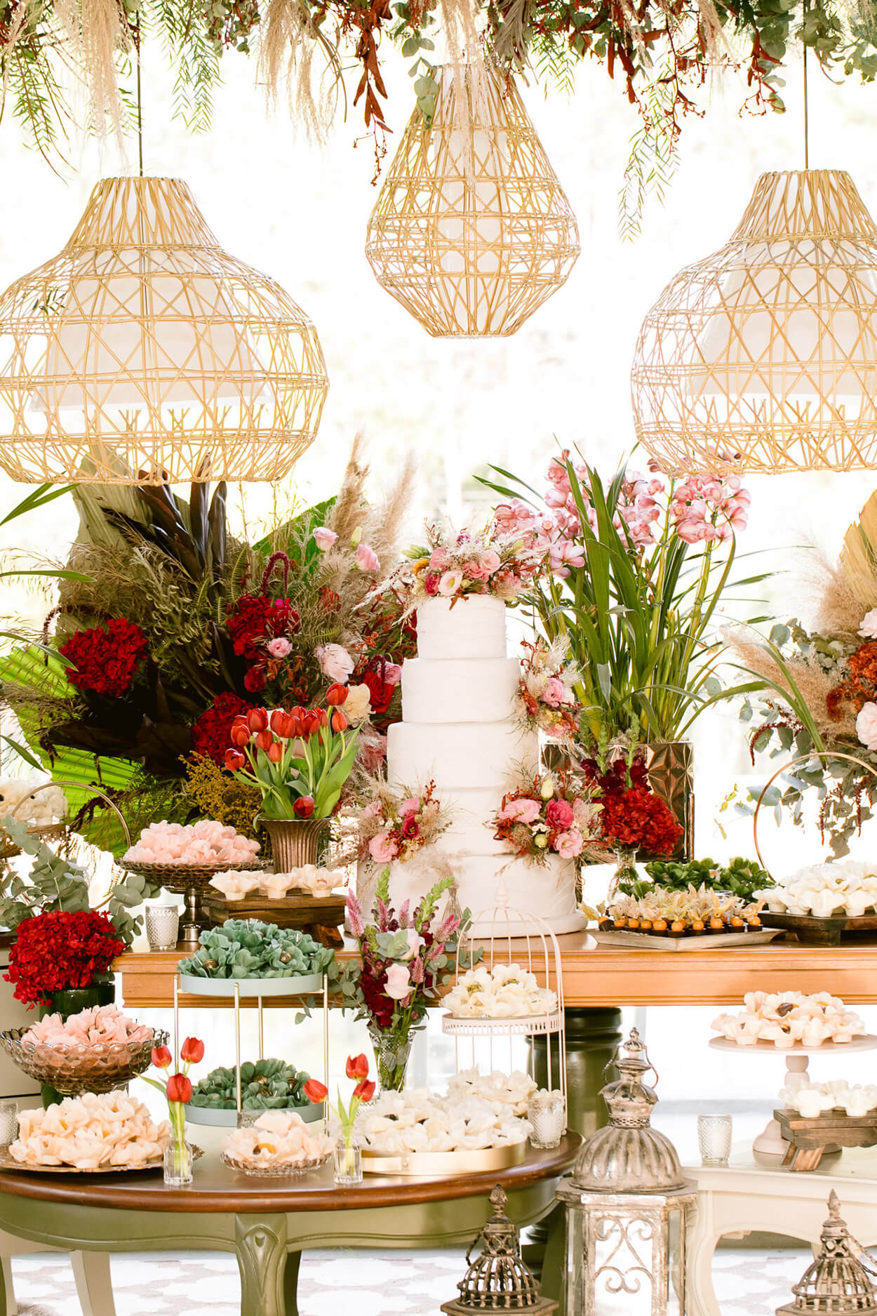 Mesa com bolo de casamento com cinco andares e vasos de flores vermelhas rosas e brancas e luminarias pendentes rusticas