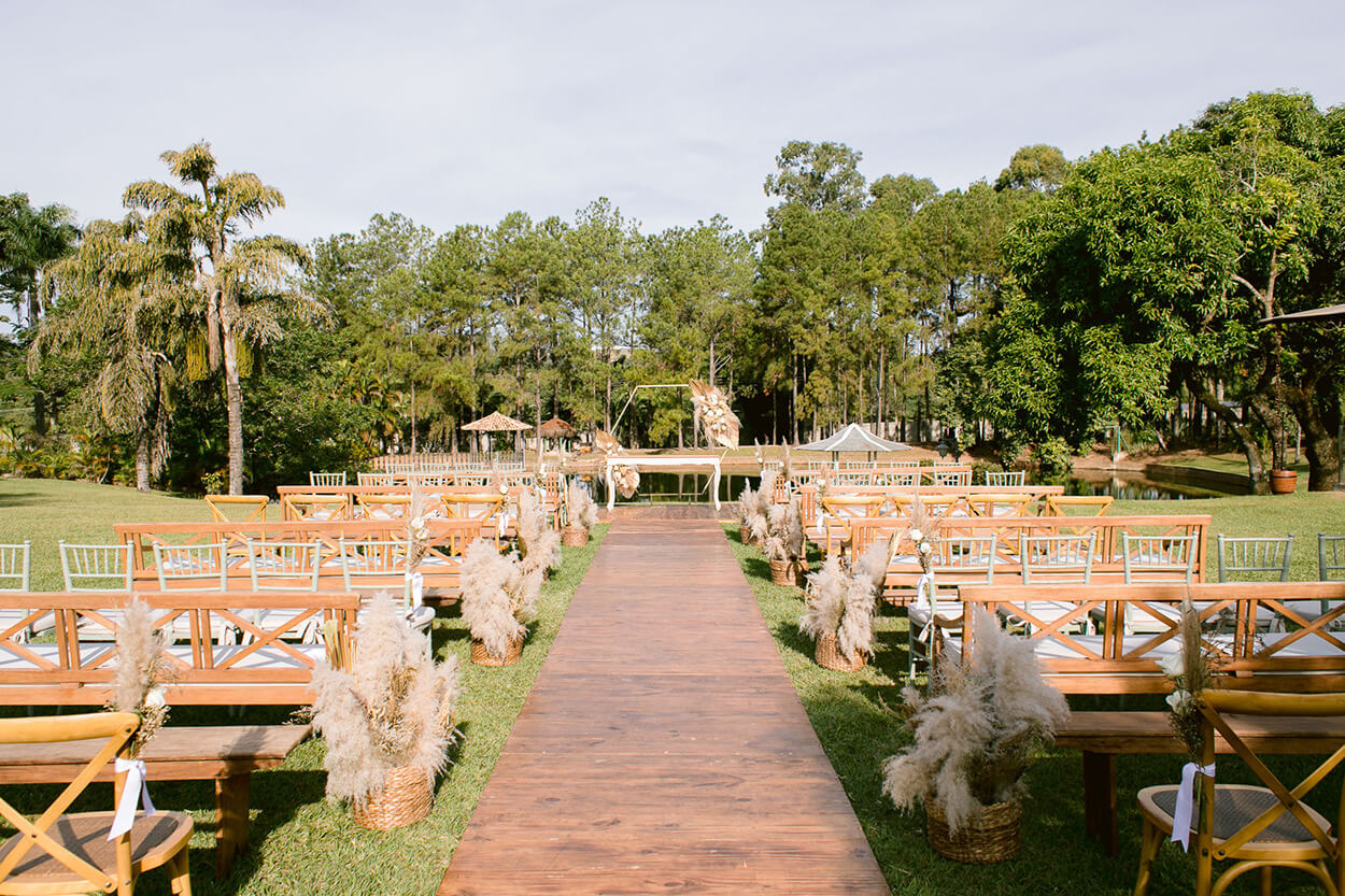 Bancos e cadeiras de madeira clara e vasos com capim dos pampas no jardim de frente para o lago para realizar casamento 